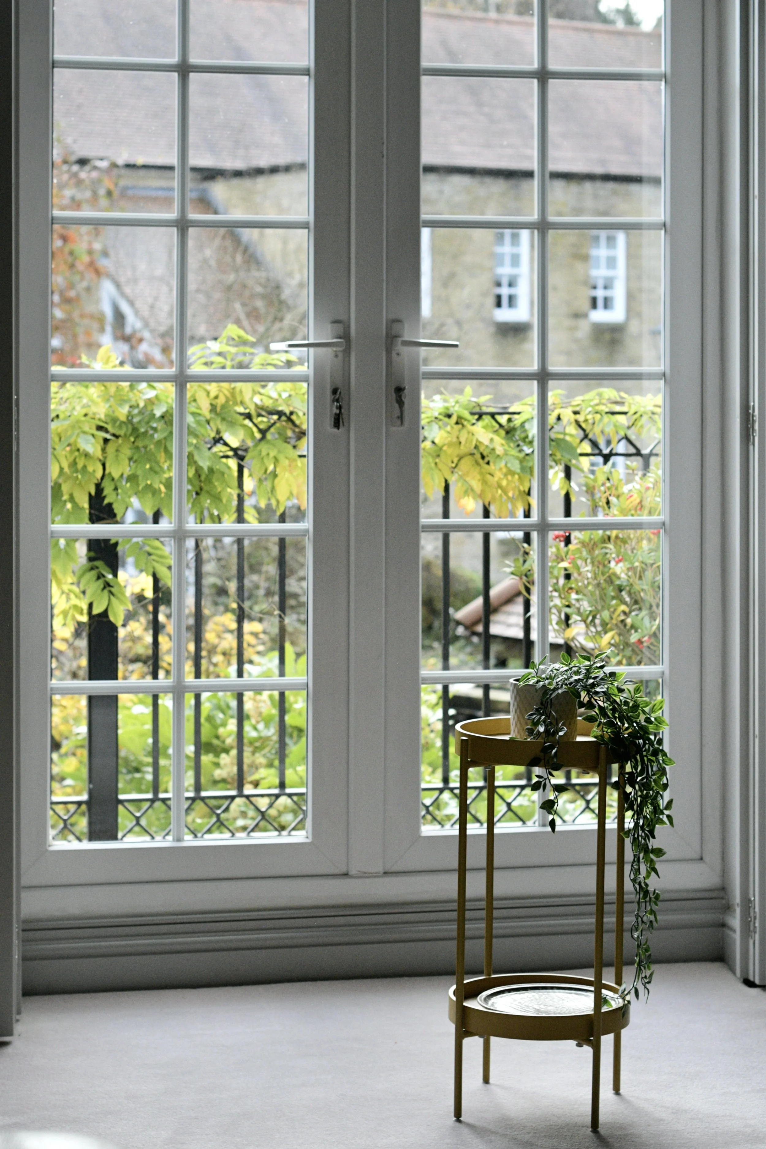 A pair of white-framed glass doors leading to an outdoor balcony with green foliage, an old stone building in the background, and a small table with potted plants inside.