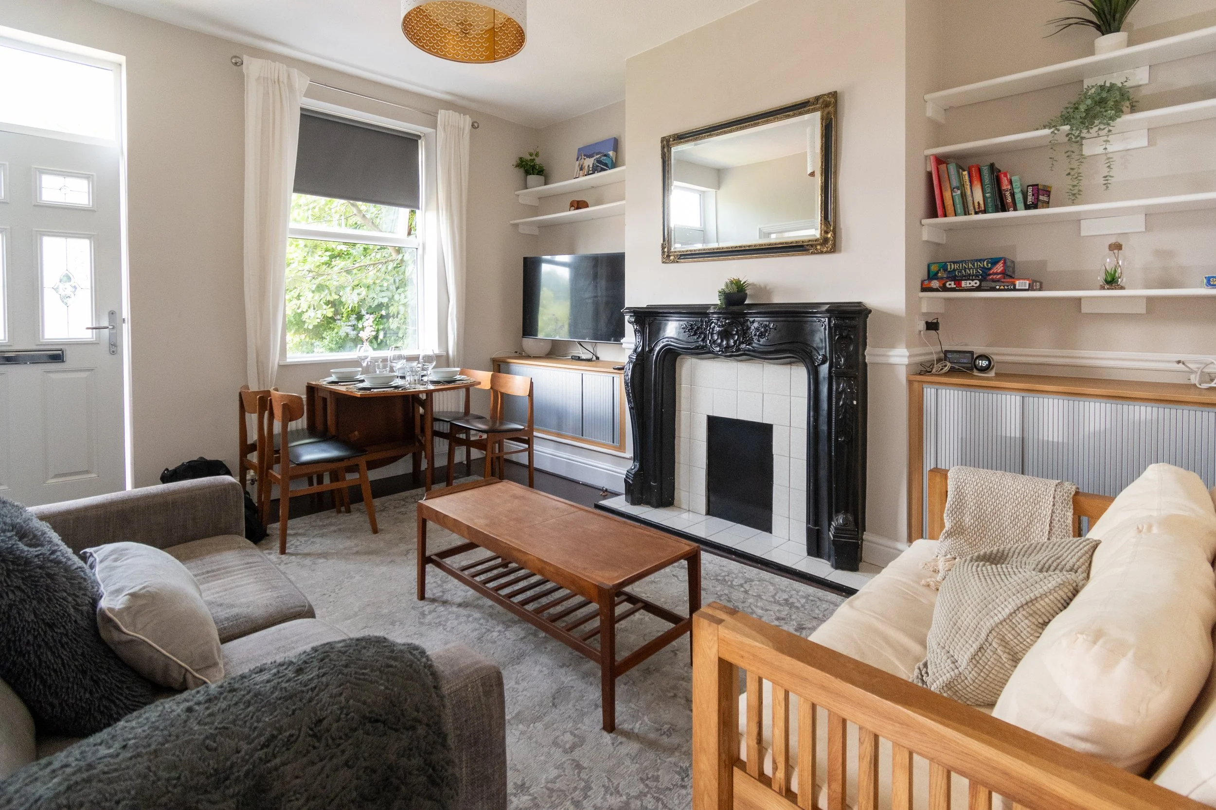 A cozy living room with two sofas, a fireplace, a wall-mounted TV, a dining table set for four, and shelves with books and decorations, near a window with white curtains and a ceiling light fixture.