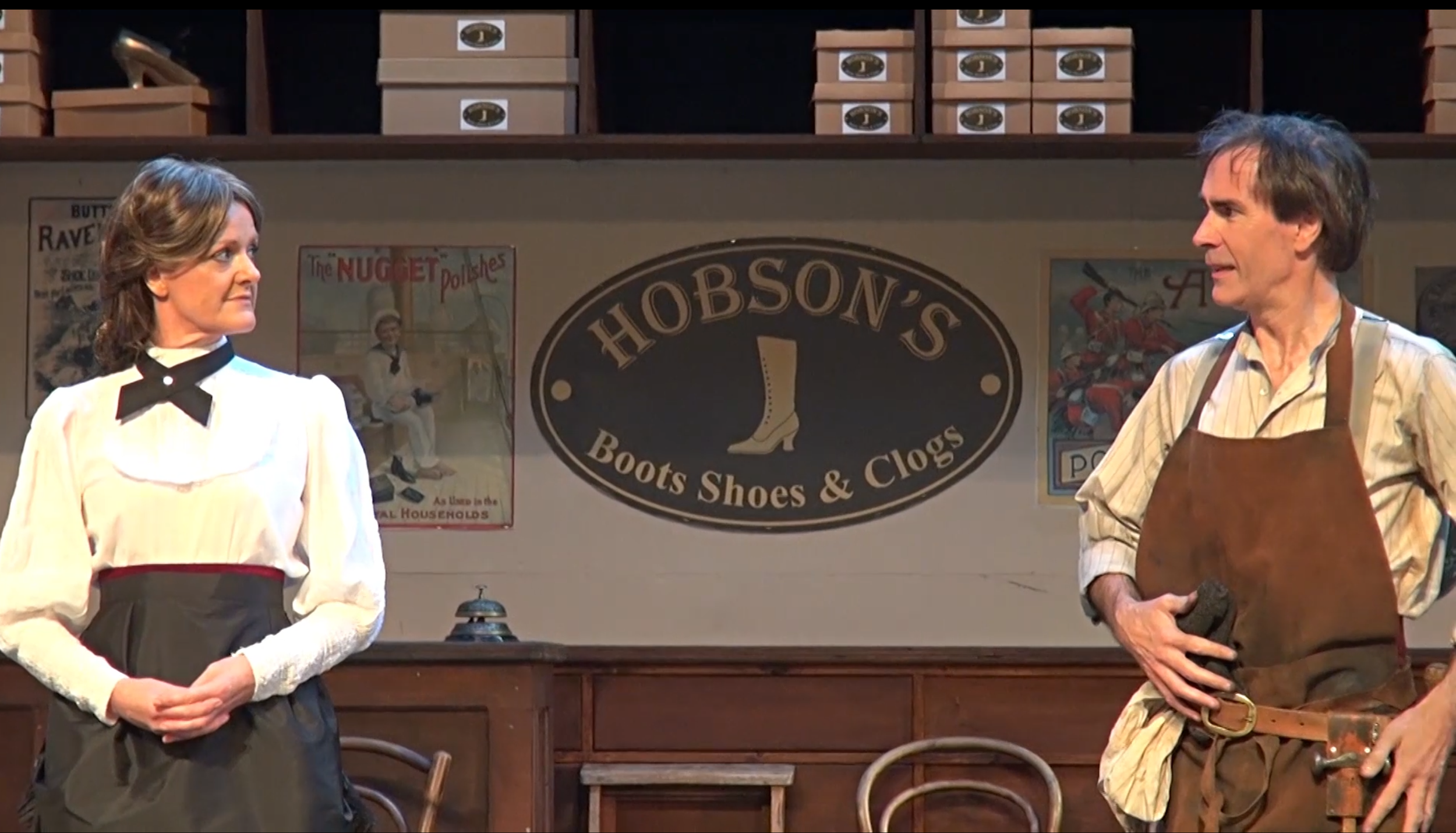 Drama scene:  Two actors in period costume onstage with a store sign reading 'Hobson's Boots Shoes & Clogs', and posters in the background.