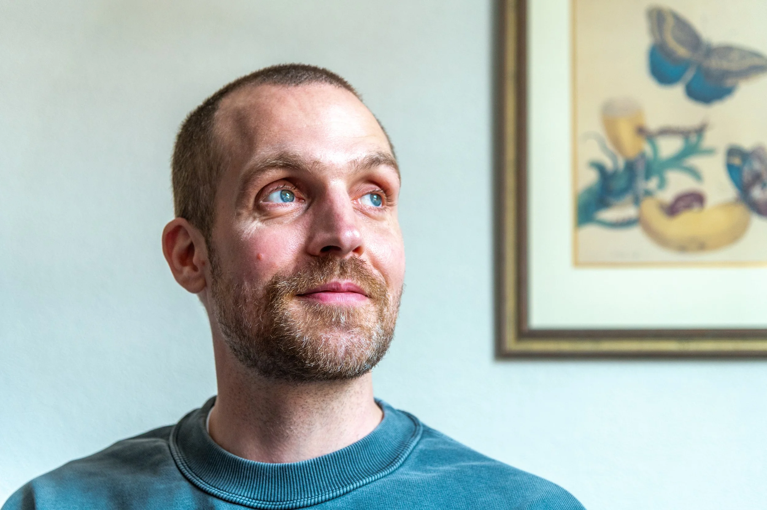 A man with light skin, short hair, and a beard looking slightly upward. He is wearing a dark-colored shirt and is standing in front of a light-colored wall with a framed artwork featuring abstract shapes and a butterfly.