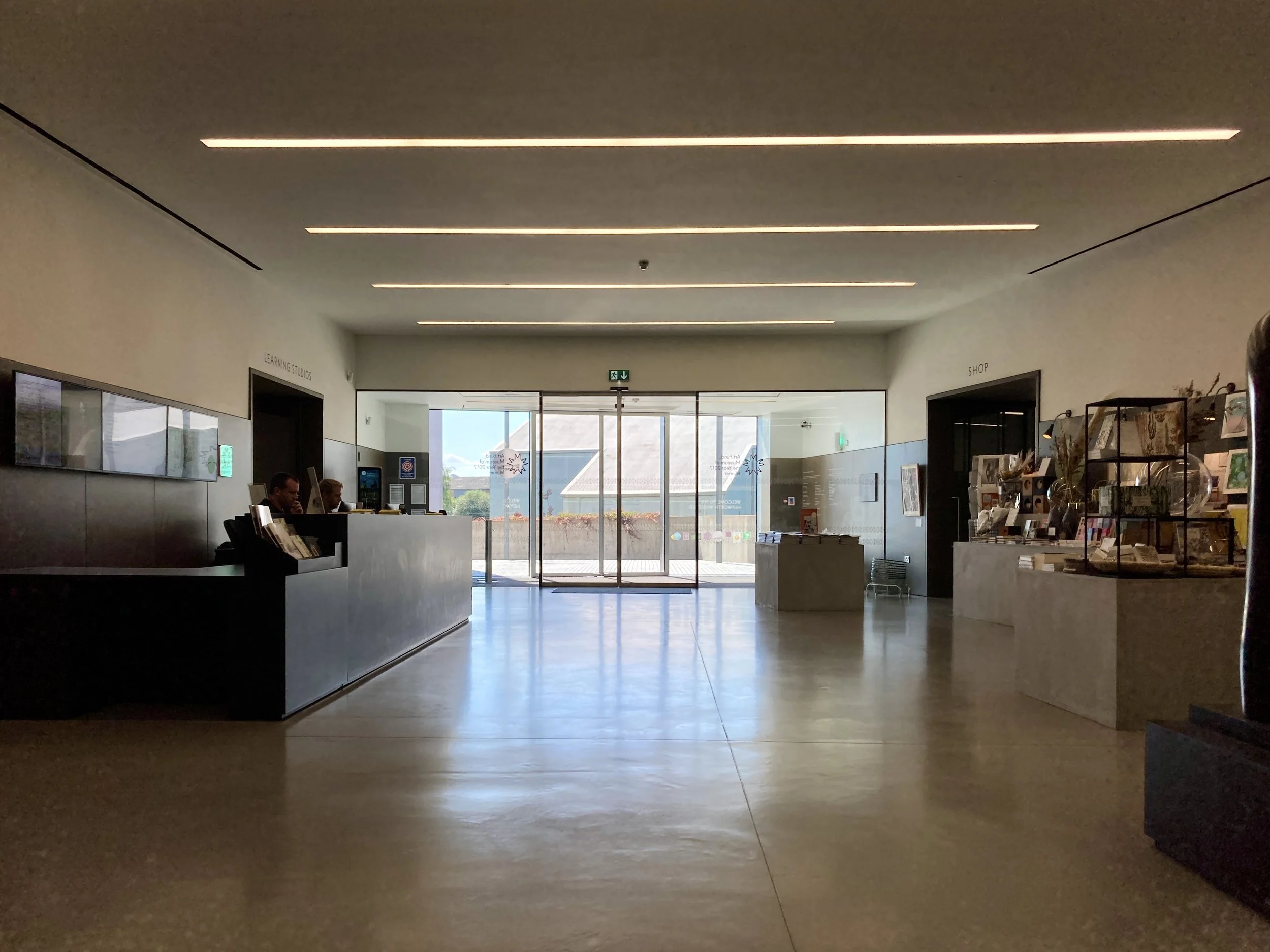 Interior of a modern building with a reception desk and large glass doors leading outside, sunlight streaming in.