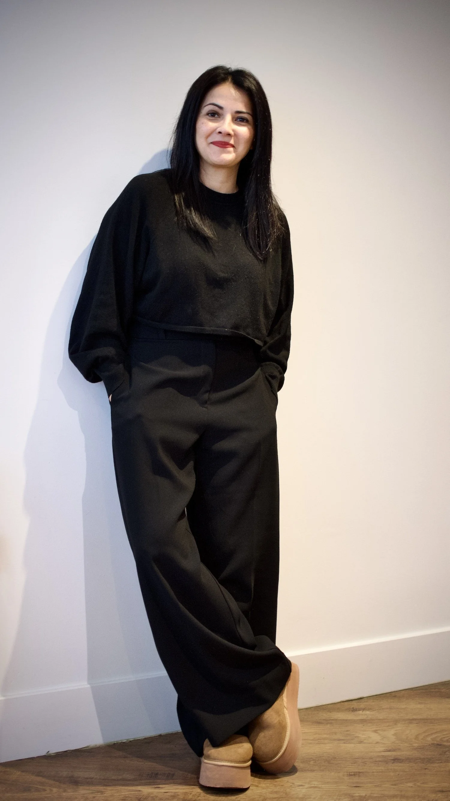 A woman with black hair and light skin, dressed in a black long-sleeve top and black pants, standing against a plain white wall, wearing tan casual shoes and smiling at the camera.