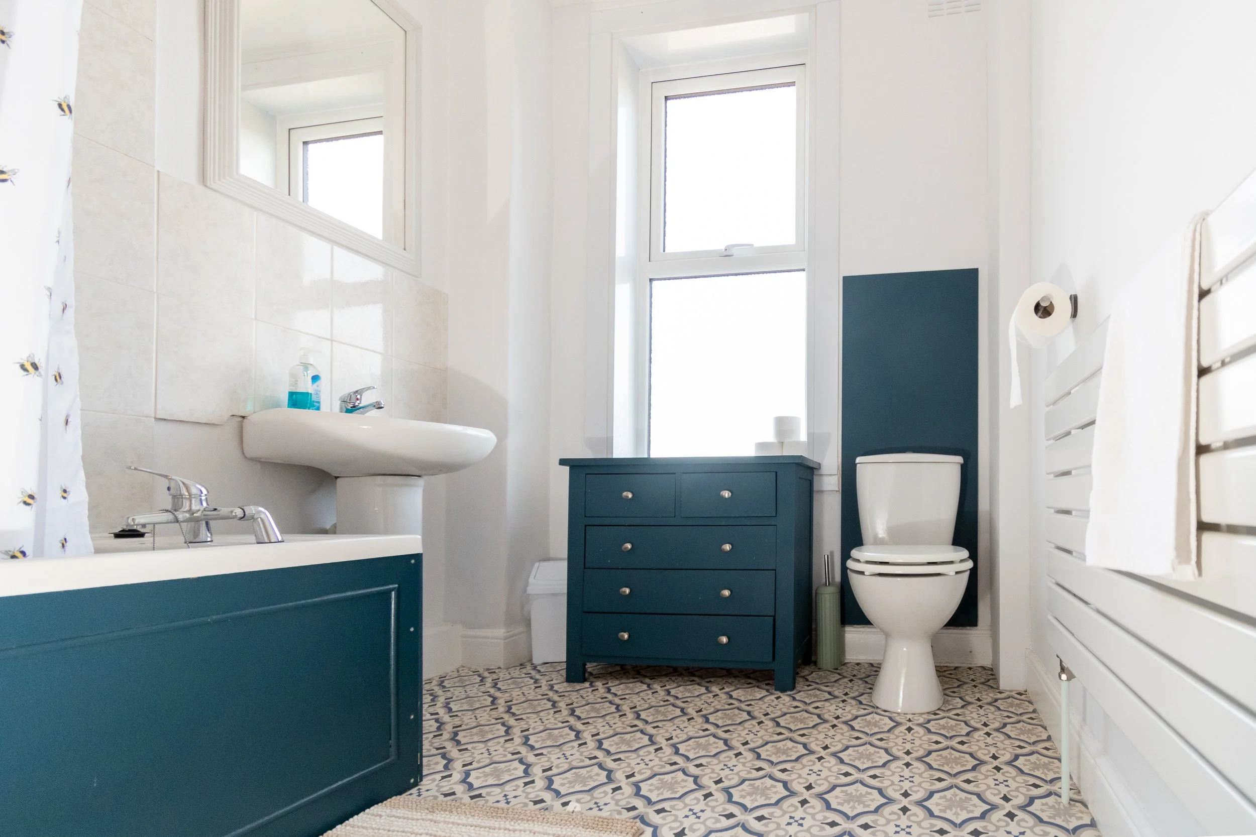 A bathroom with a white sink, blue vanity, white toilet, tiled floor, and two windows letting in natural light.
