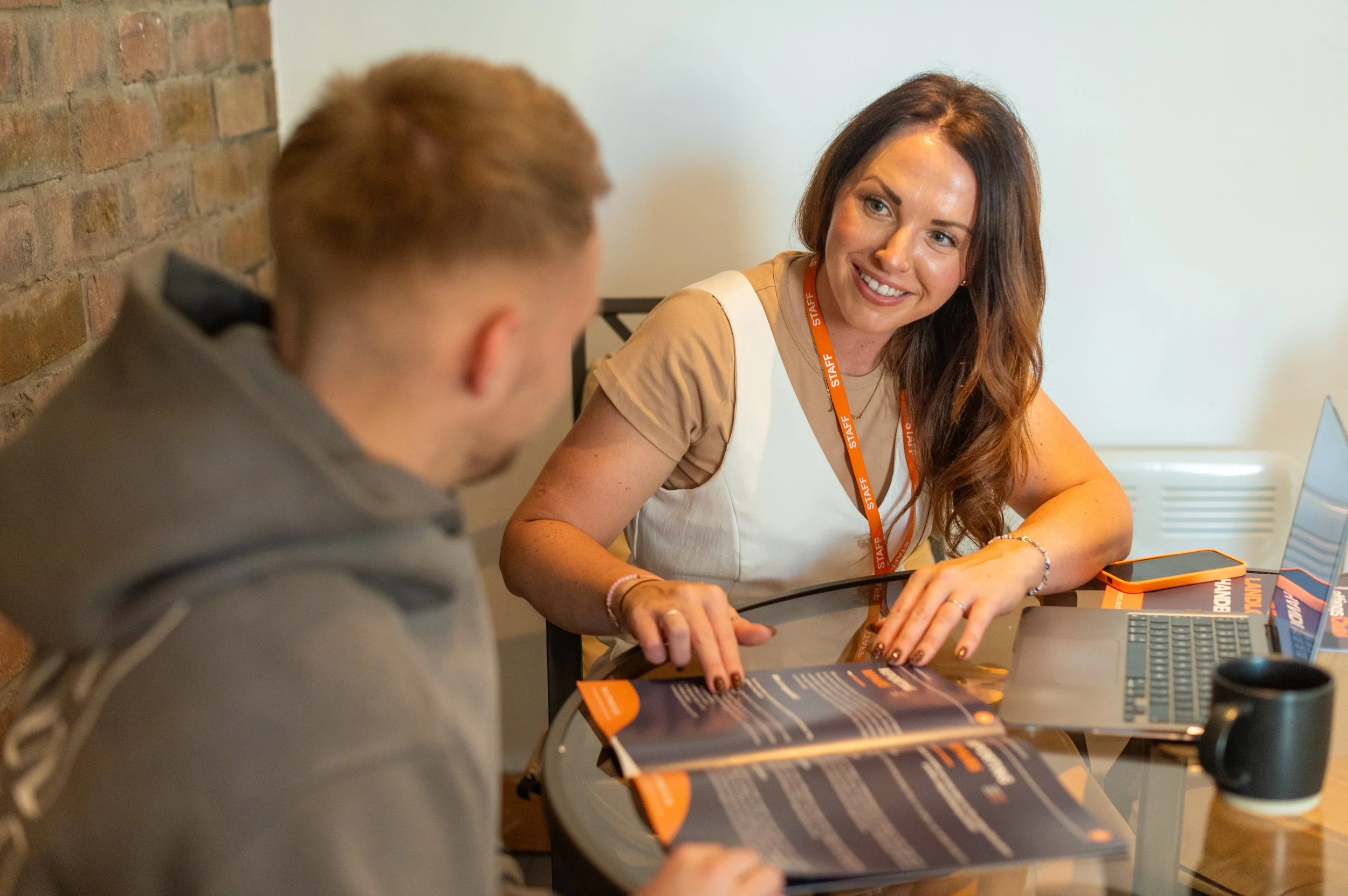 A woman with long brown hair smiling and talking to a man with short red hair, sitting at a desk with brochures, a tablet, and a laptop, in a room with a brick wall and white wall.