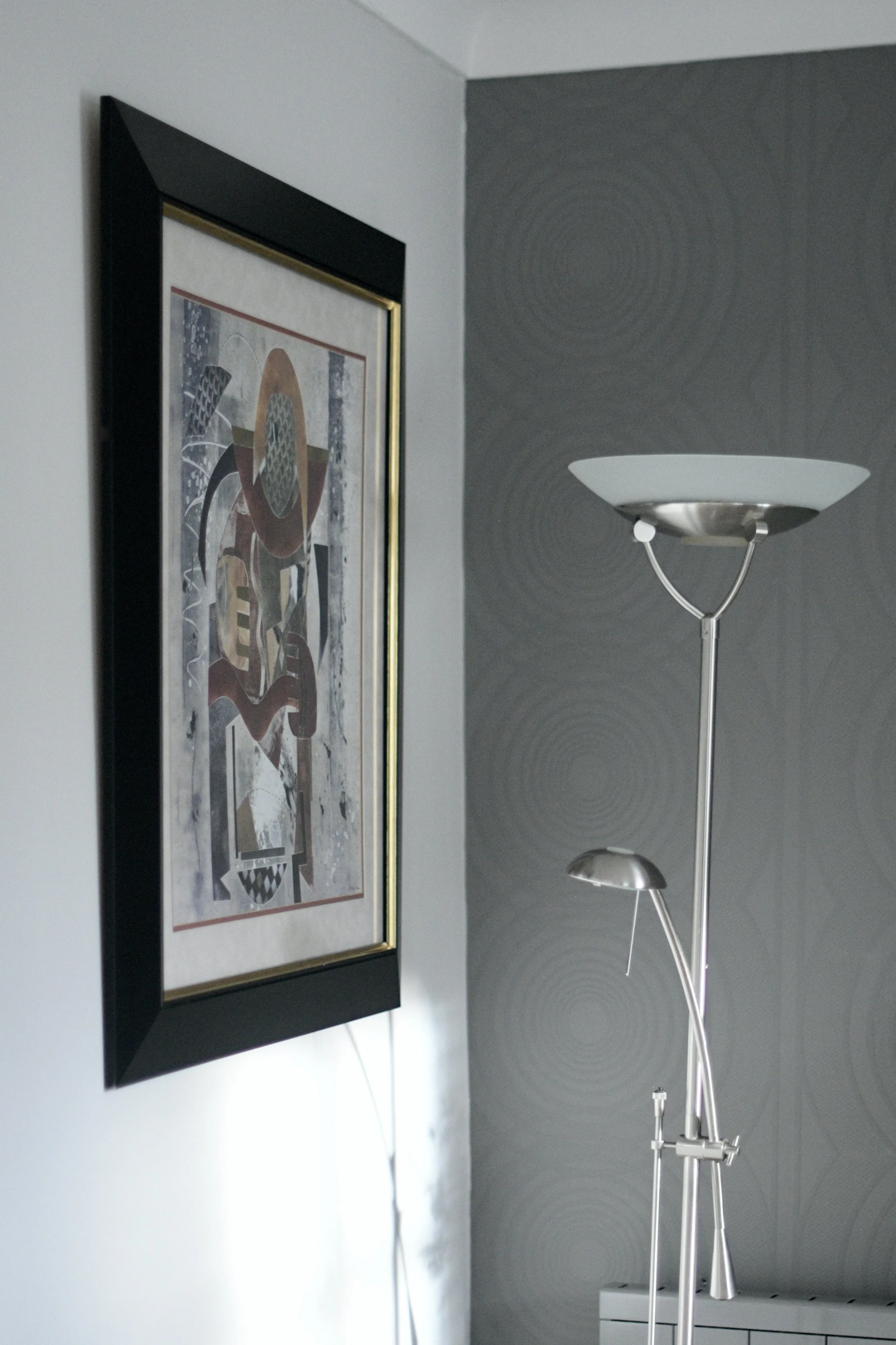 Interior shot of a room with a modern silver floor lamp with dual adjustable lamp heads, a framed abstract artwork on the wall featuring geometric shapes and earthy tones, and a gray wall with subtle circular pattern wallpaper.