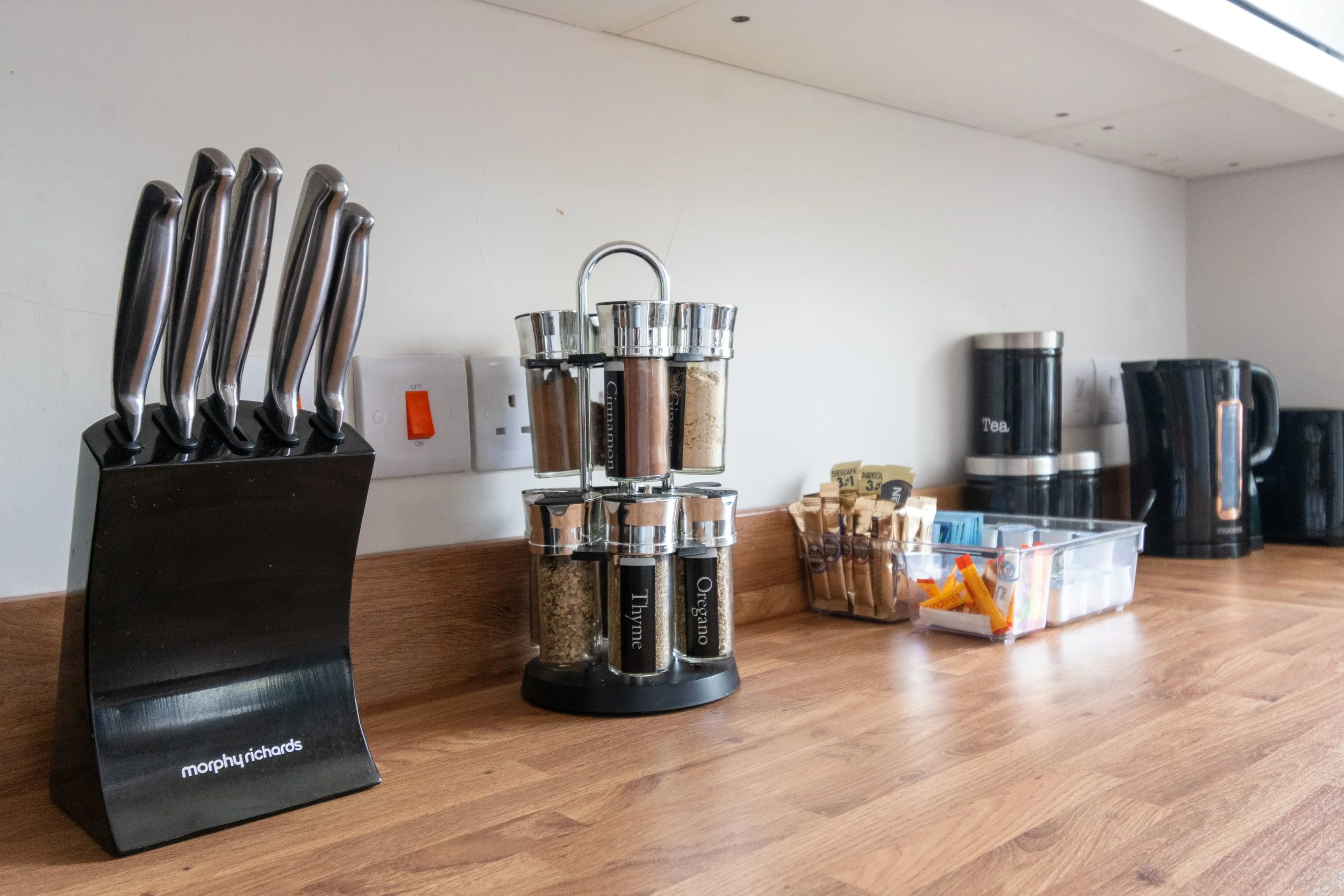A countertop with a knife set, a spice rack, tea and coffee accessories, and various small items like sugar packets and clips.