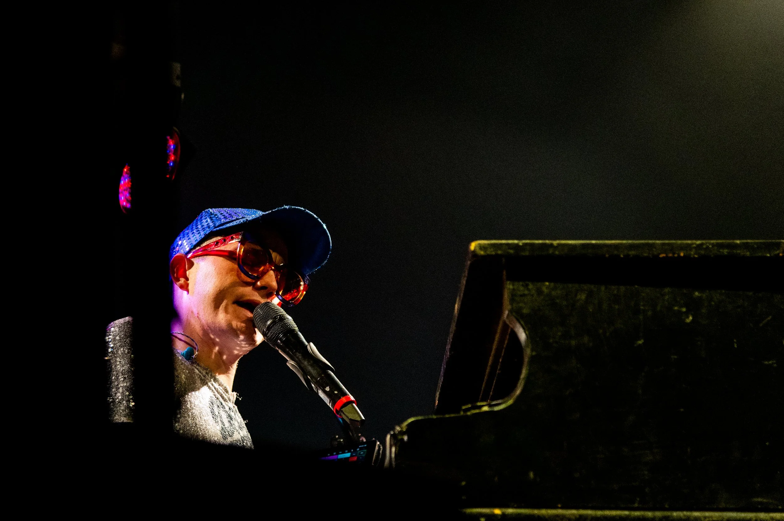 A woman singing into a microphone at night, wearing a blue cap, large red sunglasses, and a sparkly silver top, with a music stand in front of her.