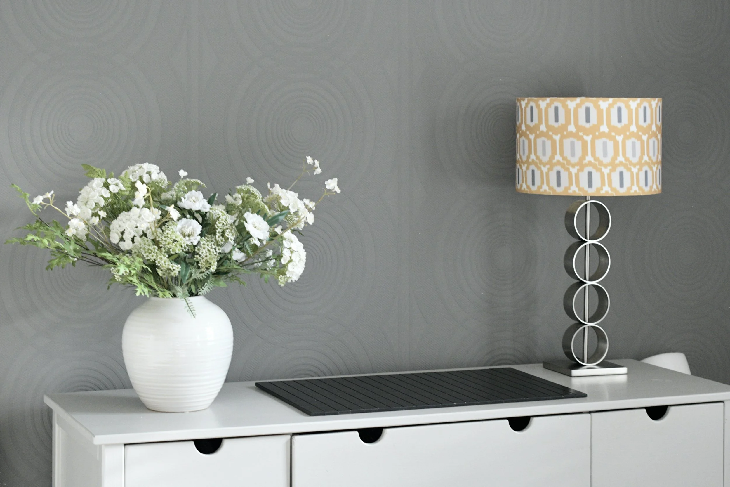 Decorative table lamp with a beige and white pattern on the lampshade, a white vase with a bouquet of white flowers, on a white sideboard with four drawers, against a textured gray wall.