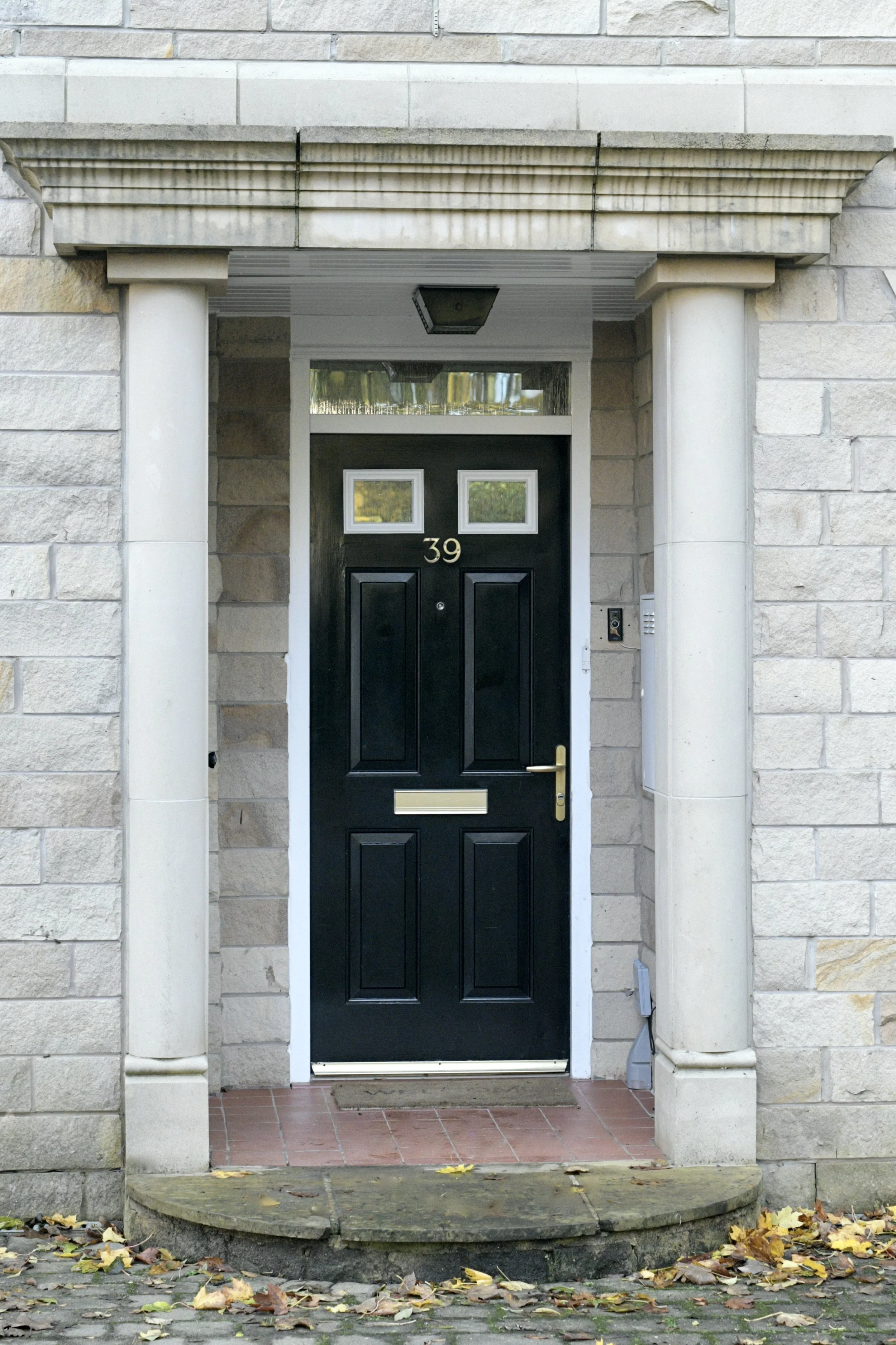 Front entrance of a house with black door, white trim, and stone columns, fallen yellow leaves on the ground.