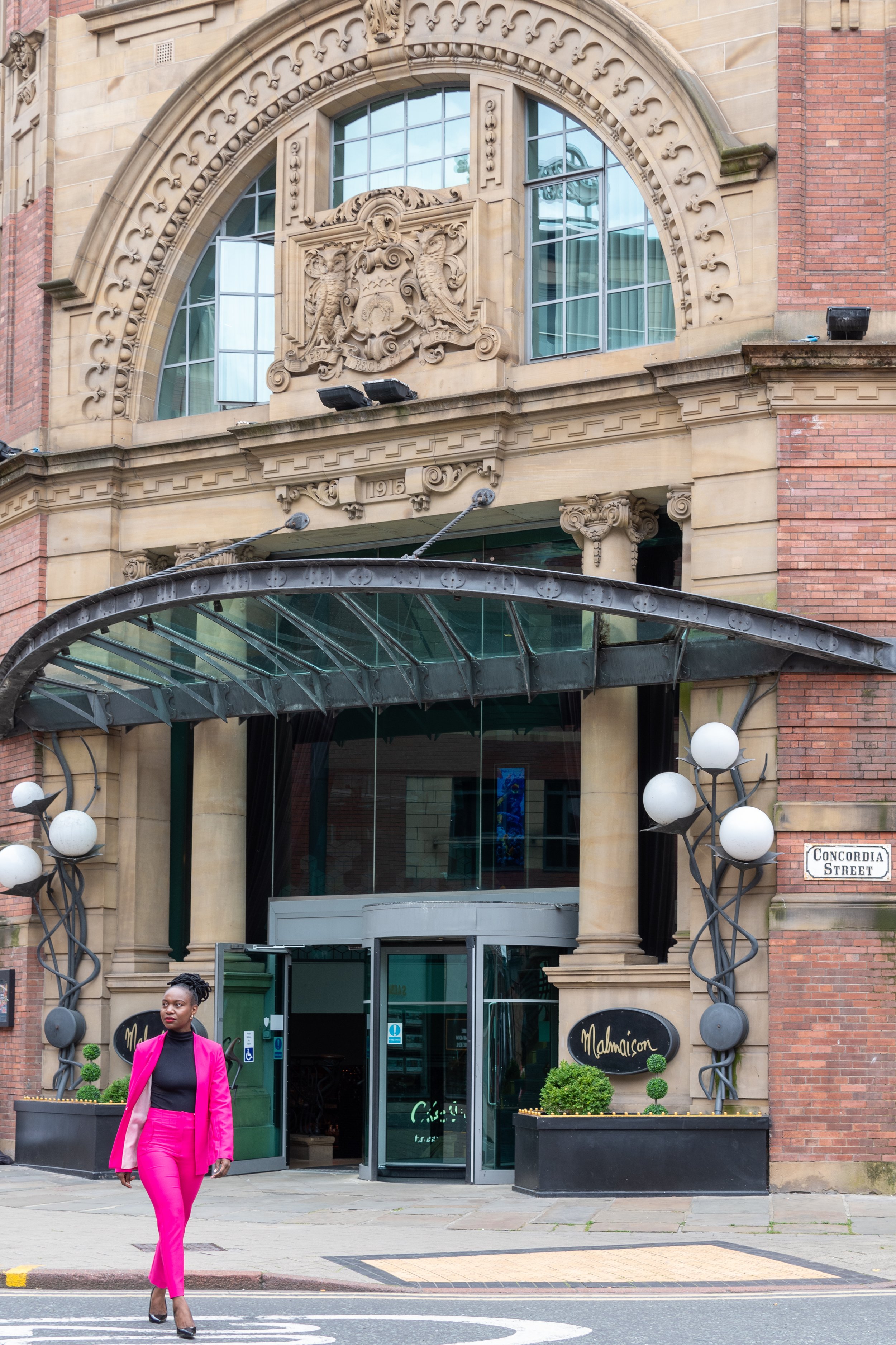 A woman dressed in a bright pink suit walking on the street in front of an ornate stone facade. The building has large windows, decorative carvings, and a sign for 'Mahamicon' at the entrance. There are planters with small, sculpted bushes and a street sign that says 'Concordia Street'.