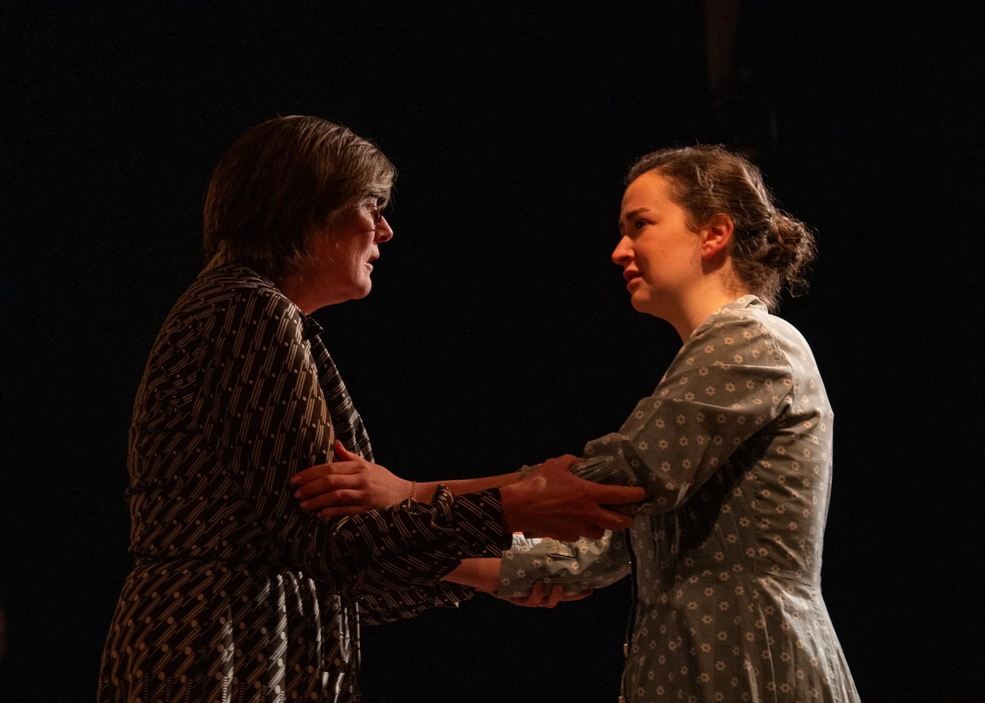 Drama scene: two women face each other, holding hands and gazing intensely into each other's eyes on a dark stage, suggesting an emotional moment or scene.