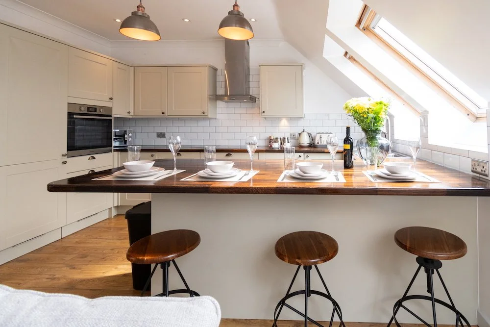 A modern kitchen with white cabinets, a wooden countertop island set for four with placemats, plates, and glasses, three wooden barstools, a glass vase with yellow flowers, a bottle of wine, and a window on a sloped ceiling.