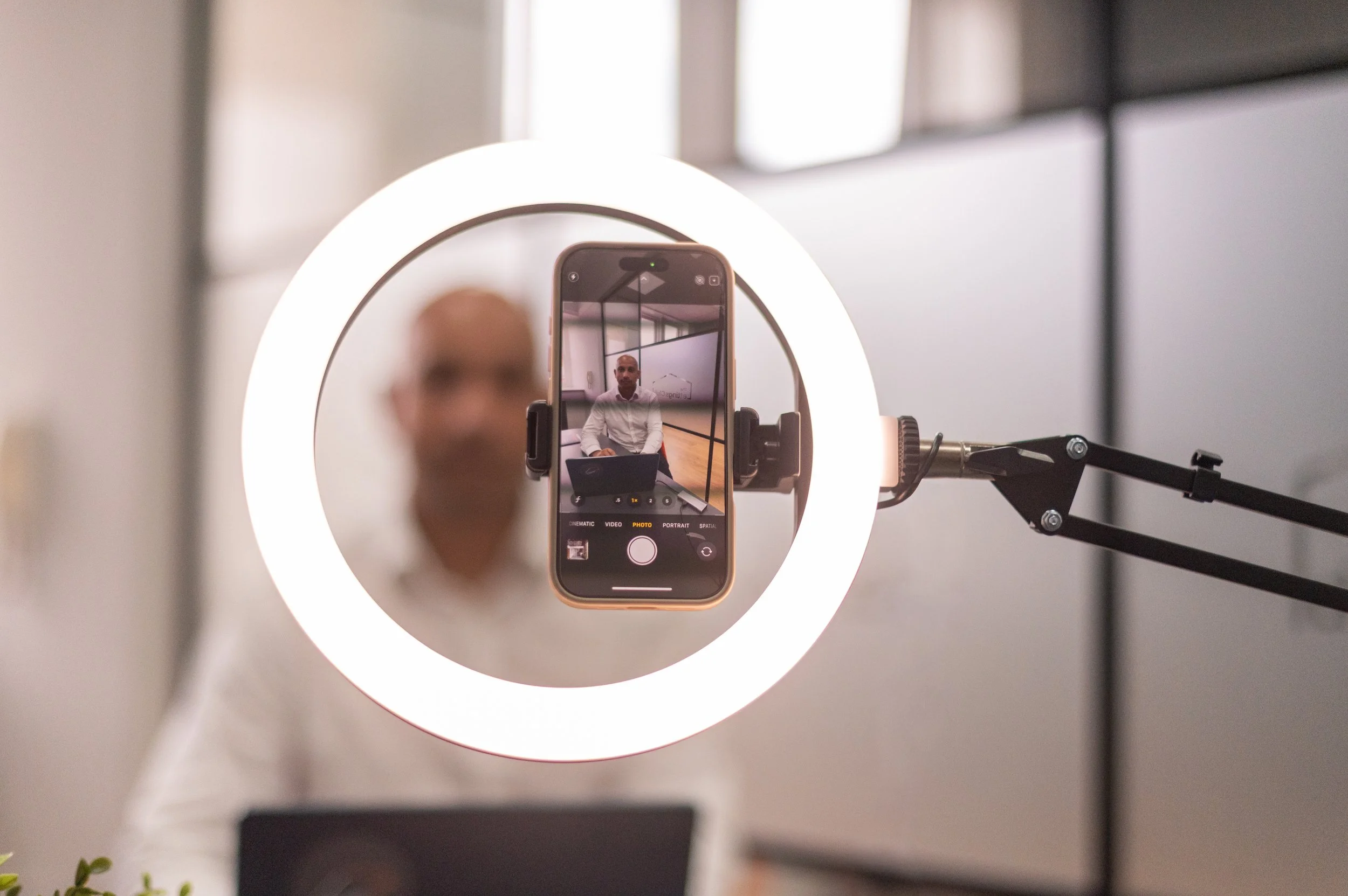 A smartphone mounted on a ring light captures a photo of a bald man in a white shirt seated at a table in a modern indoor setting.