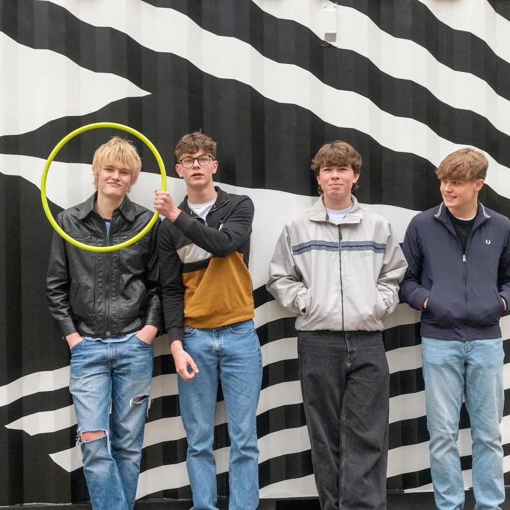Four teenage boys standing in front of a black and white striped mural. One boy is holding a yellow hula hoop framing the other's head.