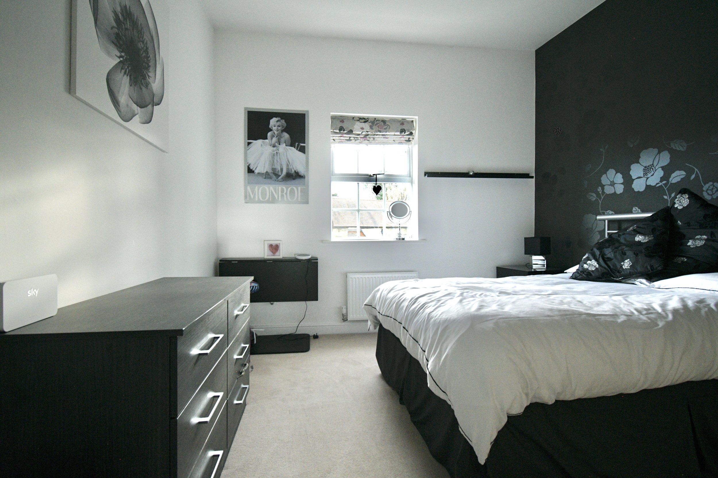 A bedroom with a white wall and a dark accent wall with floral decals. There is a window with floral blinds, a black-and-white photograph of Marilyn Monroe, and black furniture including a dresser, bedside table, and a bed with black and white bedding. Decorations include heart-shaped items and a small framed picture.