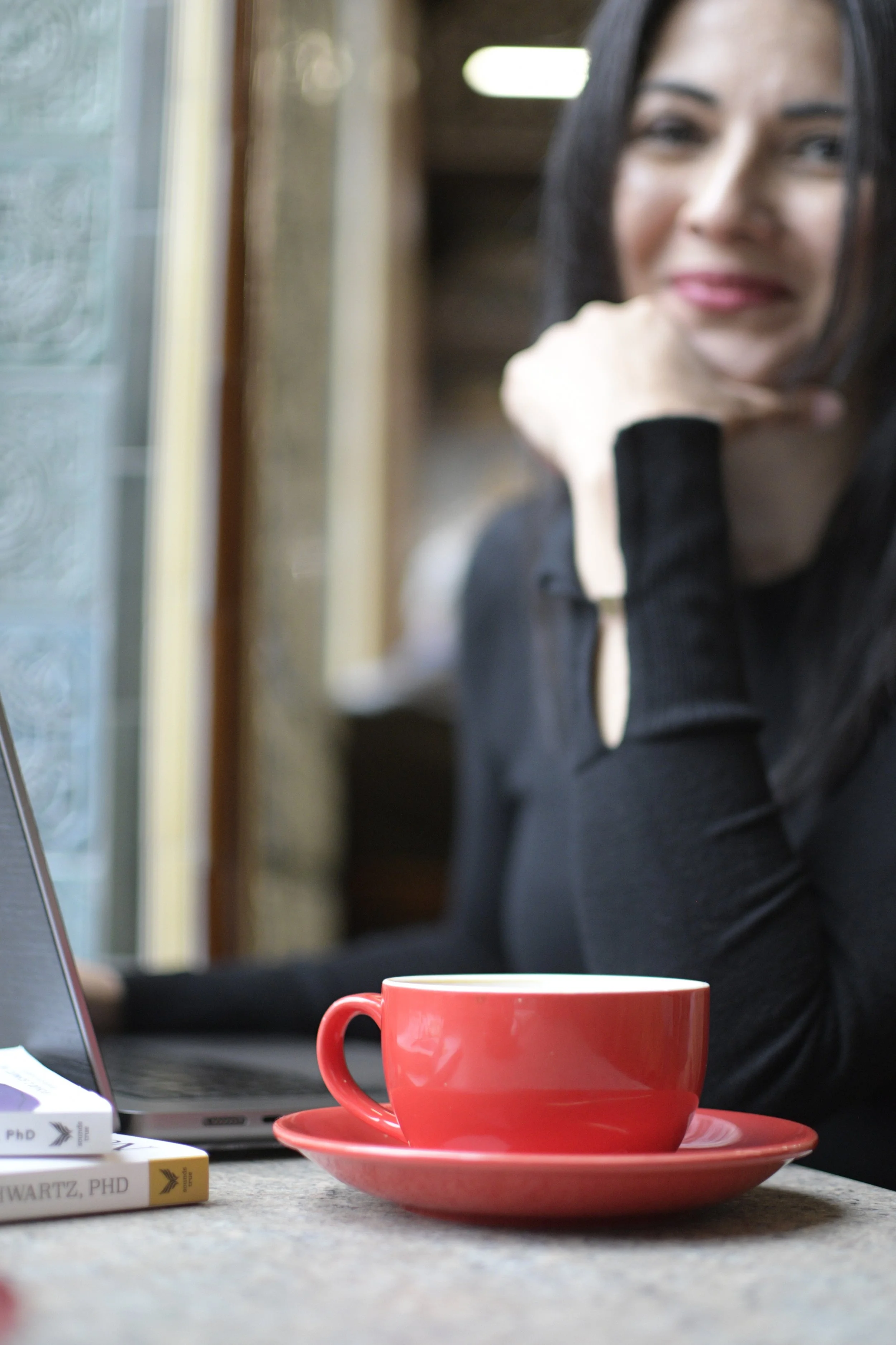 A woman with dark hair and fair skin sitting at a table, soft-focus and smiling, with a red coffee mug and saucer in front of her. She is wearing a black top and is resting her chin on her hand.