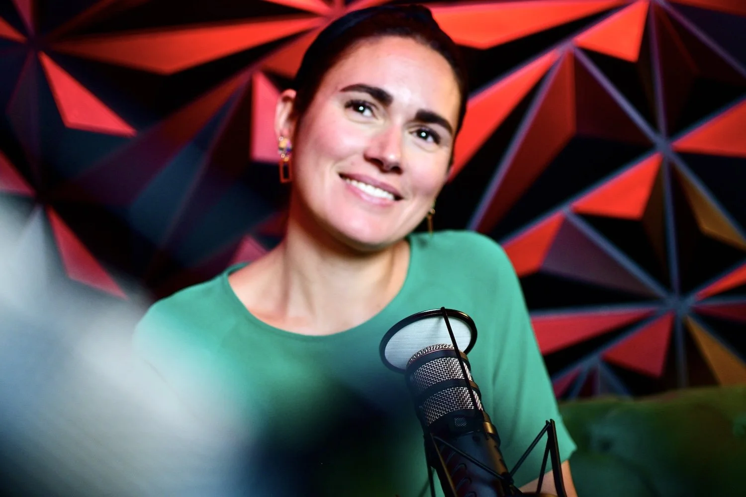 A woman with short dark hair, wearing earrings and a green top, smiling in front of a colorful geometric background, sitting in front of a microphone.