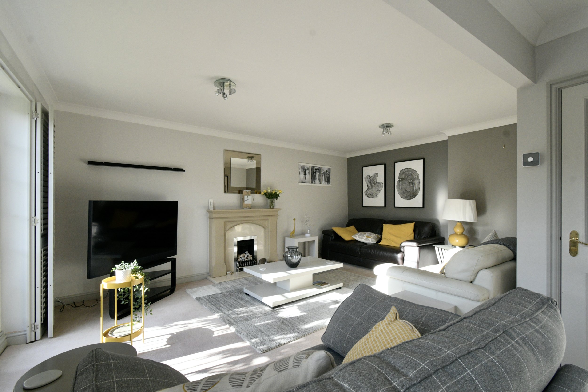 Living room with white and gray walls, a black sofa with yellow pillows, a white coffee table, a gray armchair with white cushions, a yellow side table with a plant, a television, a fireplace with decorative items, framed black and white artwork, and a white floor lamp.
