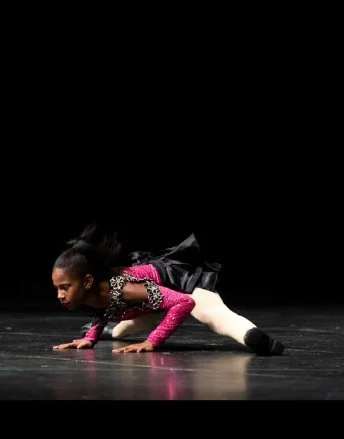 A woman performing a dance move on stage, balancing on her hands with her legs extended behind her, wearing a pink and black costume with white tights.