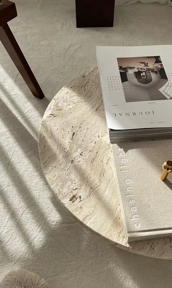 Close-up of a marble coffee table with a stack of magazines on top, a small gold object partially visible, and sunlight casting shadows on a light-colored carpet.