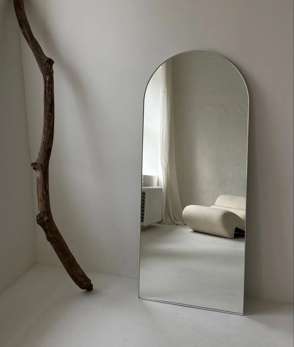 A tall, curved mirror resting on the floor, reflecting a white, modern sofa and a window with white curtains in a minimalistic room with white walls and a light-colored floor.