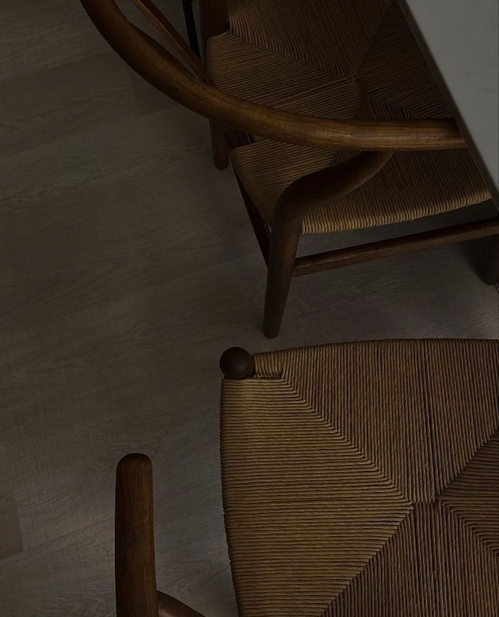 Part of a wooden dining table and two matching wooden chairs with woven seats and backrests, viewed from above in a dimly lit room.