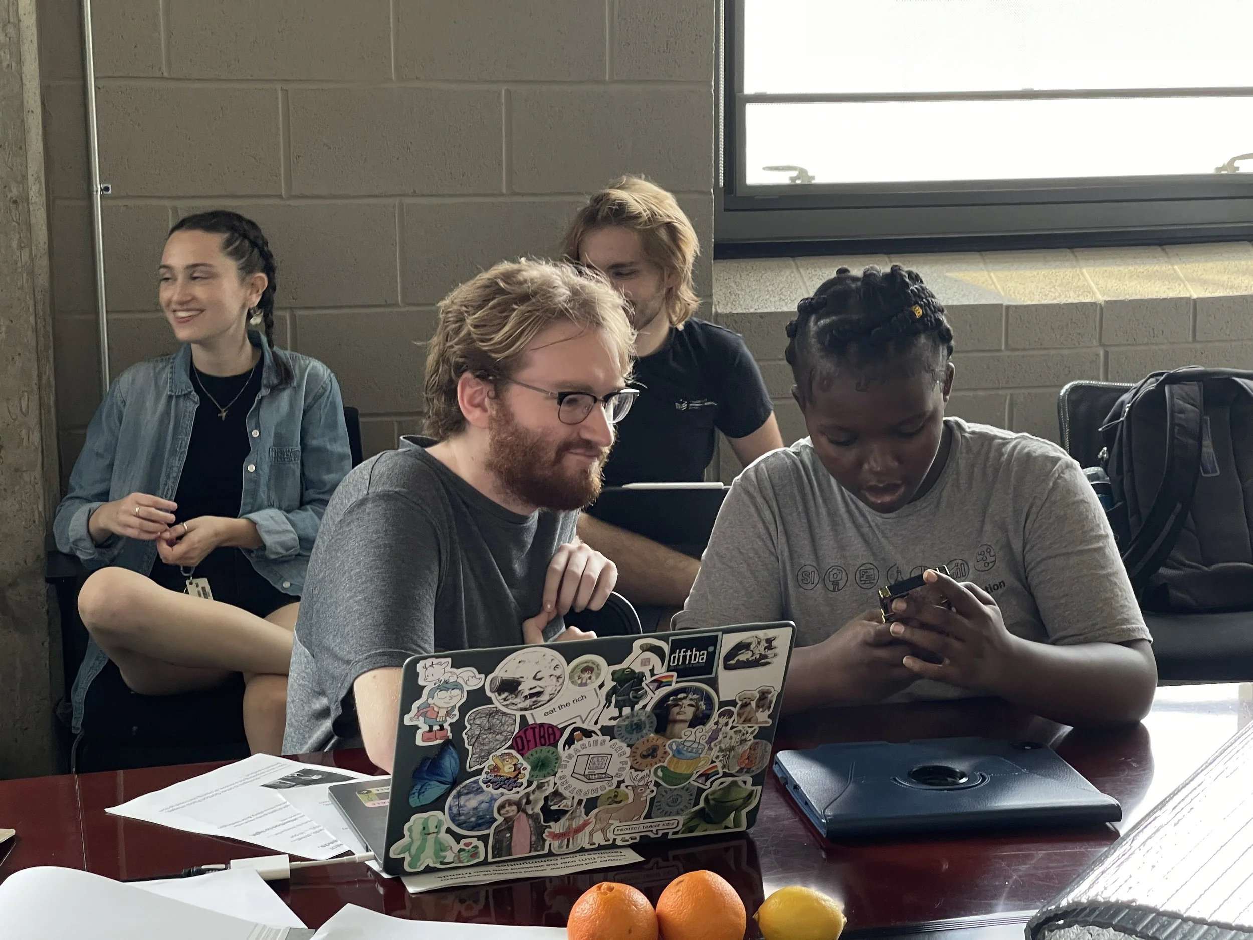 A group of four people sitting at a table, engaged with their devices, with a laptop covered in stickers, some papers, and oranges and lemons on the table. They are in a classroom with a large window and concrete wall.