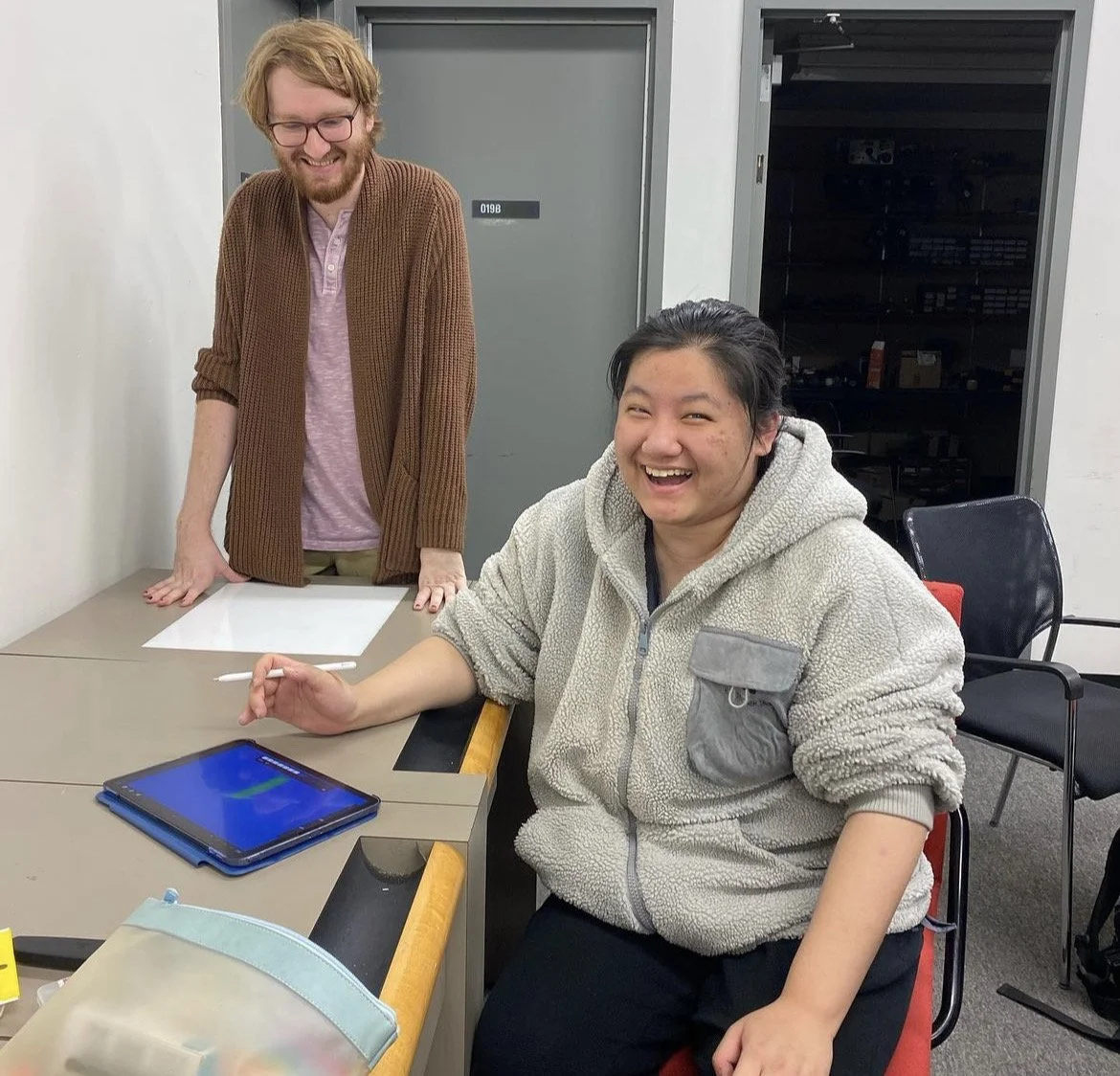 Evan Bode and an animation student in a classroom at an animation workshop, sitting at the table with a tablet and a stylus, smiling and looking towards the camera, while the teacher stands and leans on the table, both looking happy and engaged.