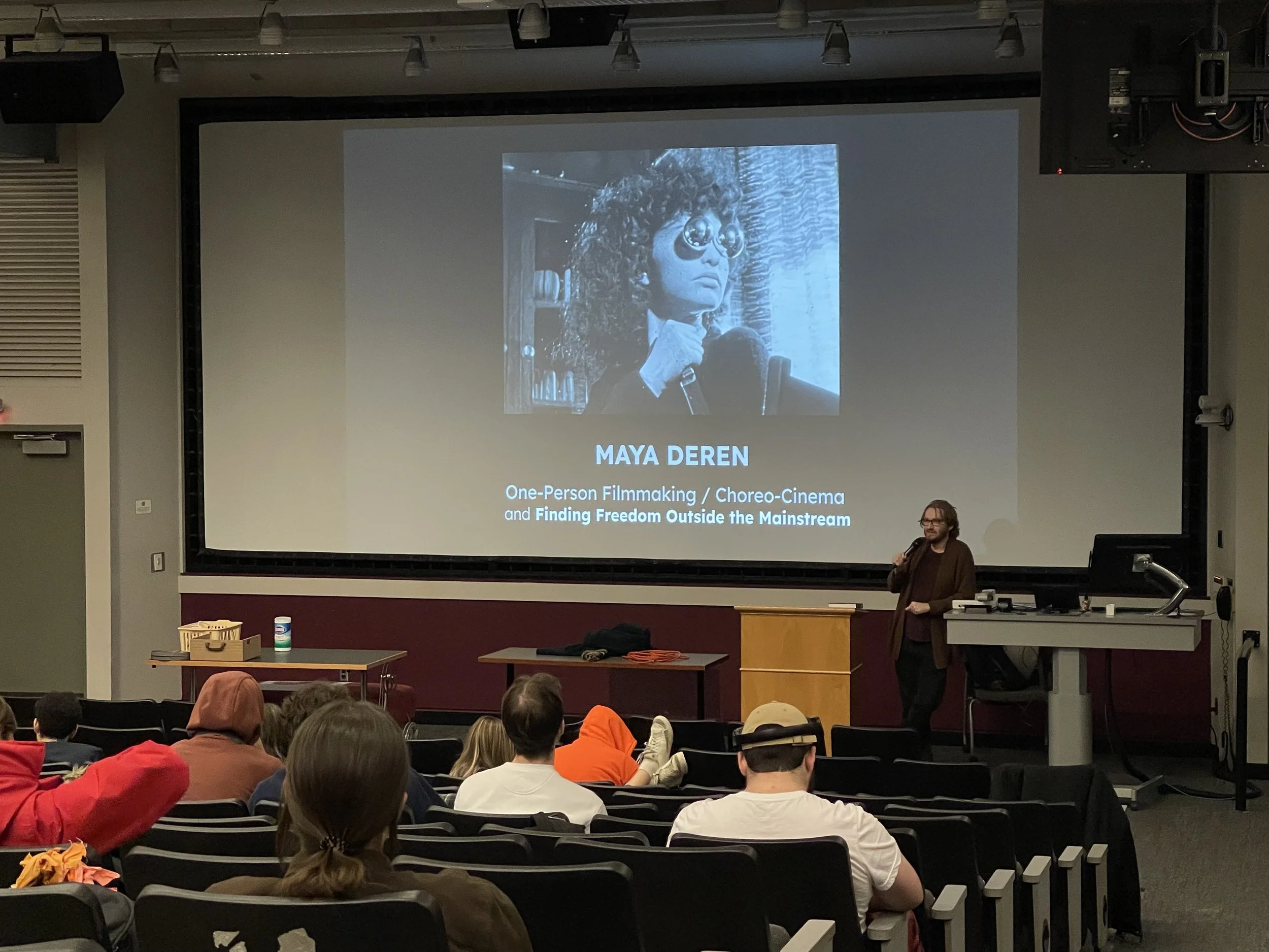 Presentation in a lecture hall about Maya Deren, featuring a large projection screen displaying her portrait, with film instructor Evan Bode standing at a podium and audience members seated, some taking notes or looking forward.