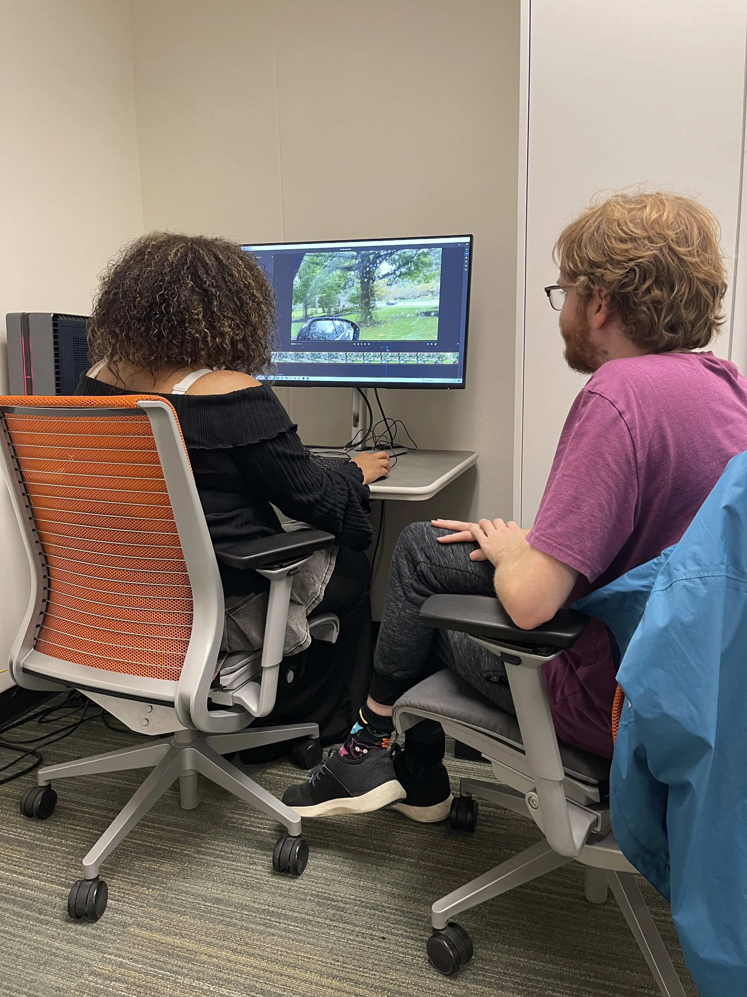 Evan Bode sits with a student in a small classroom studio, looking at a computer screen depicting a animation program with images of trees and a car.