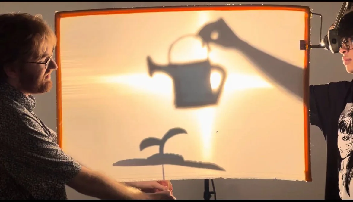 Evan Bode and one of their film students are holding a backlit screen, creating a shadow art image of a watering can pouring water onto a sprouting plant.