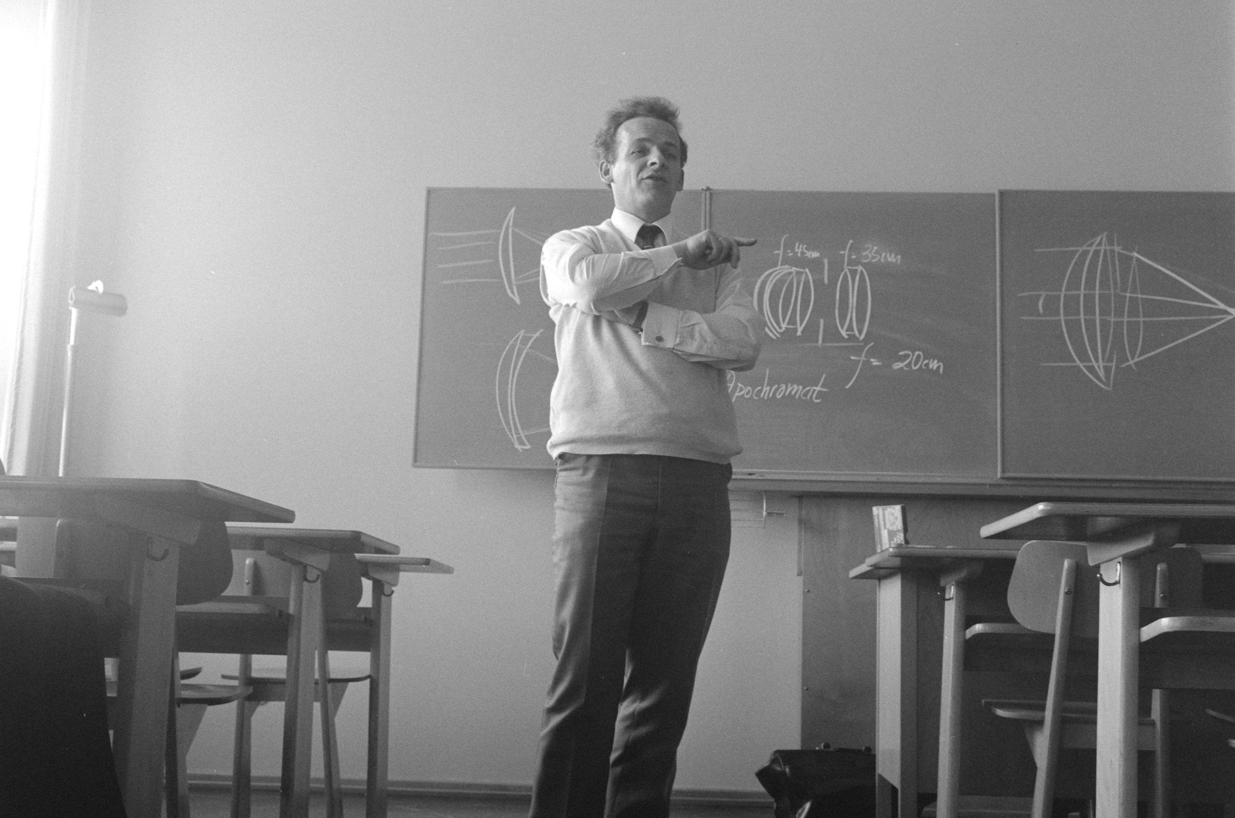A man standing in a classroom in front of a chalkboard with lens diagrams.