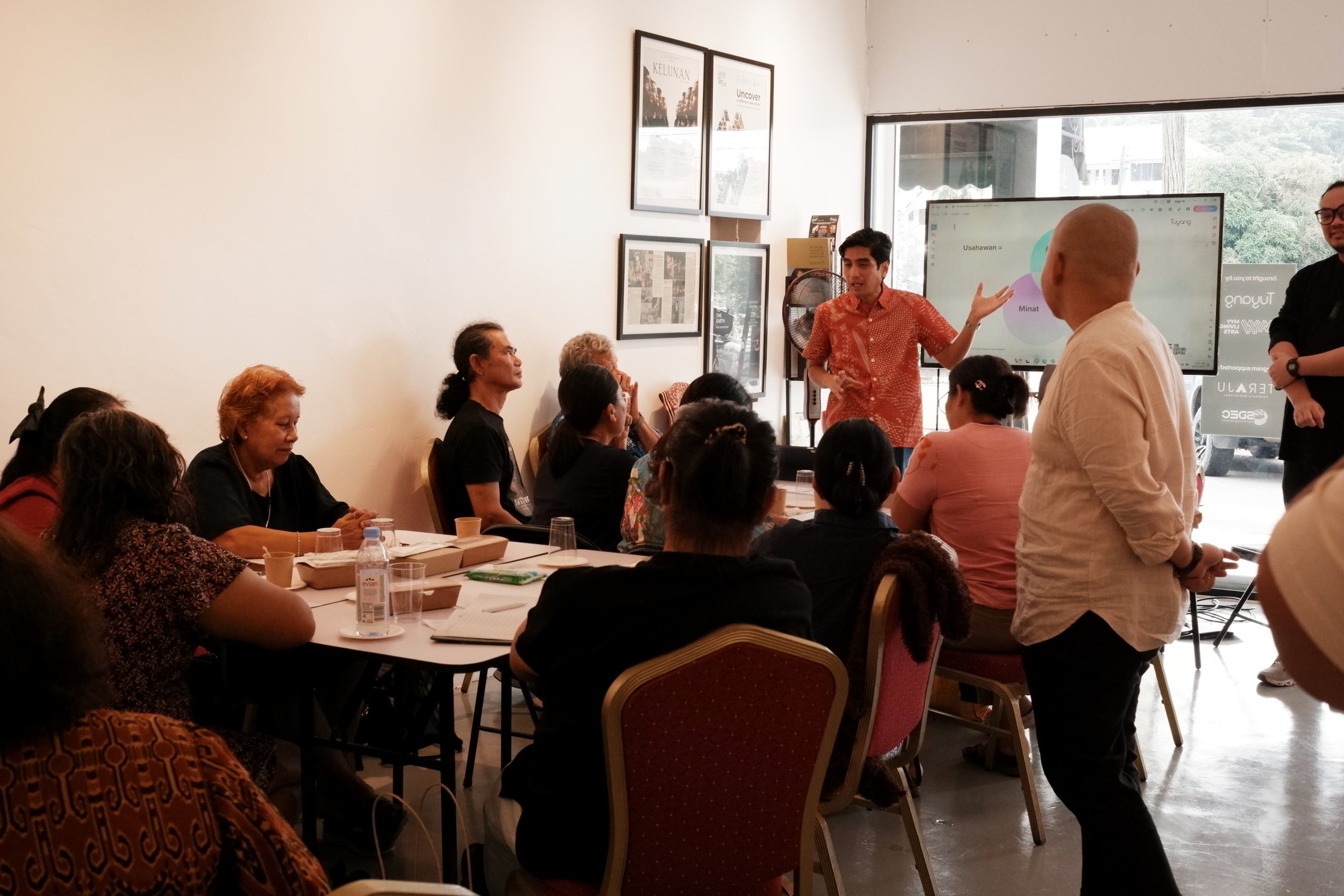 A man in a red patterned shirt giving a presentation to a group of diverse women sitting at tables in a brightly lit room, with a large screen showing a colorful diagram behind him.