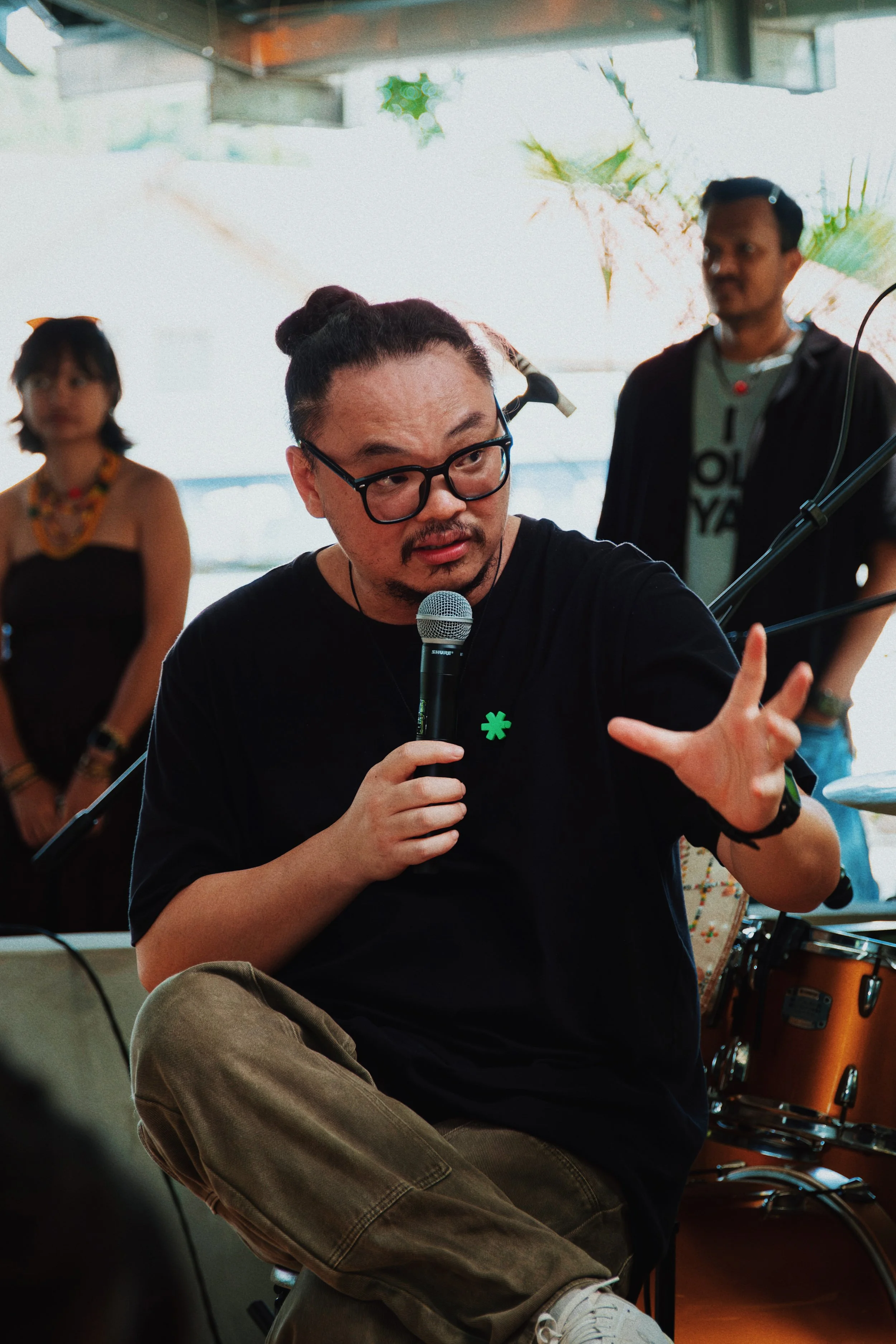 A man with glasses and a man bun is sitting cross-legged, holding a microphone, and making a hand gesture during a discussion or presentation. There are two people in the background, one woman and one man, with musical instruments and equipment aroun