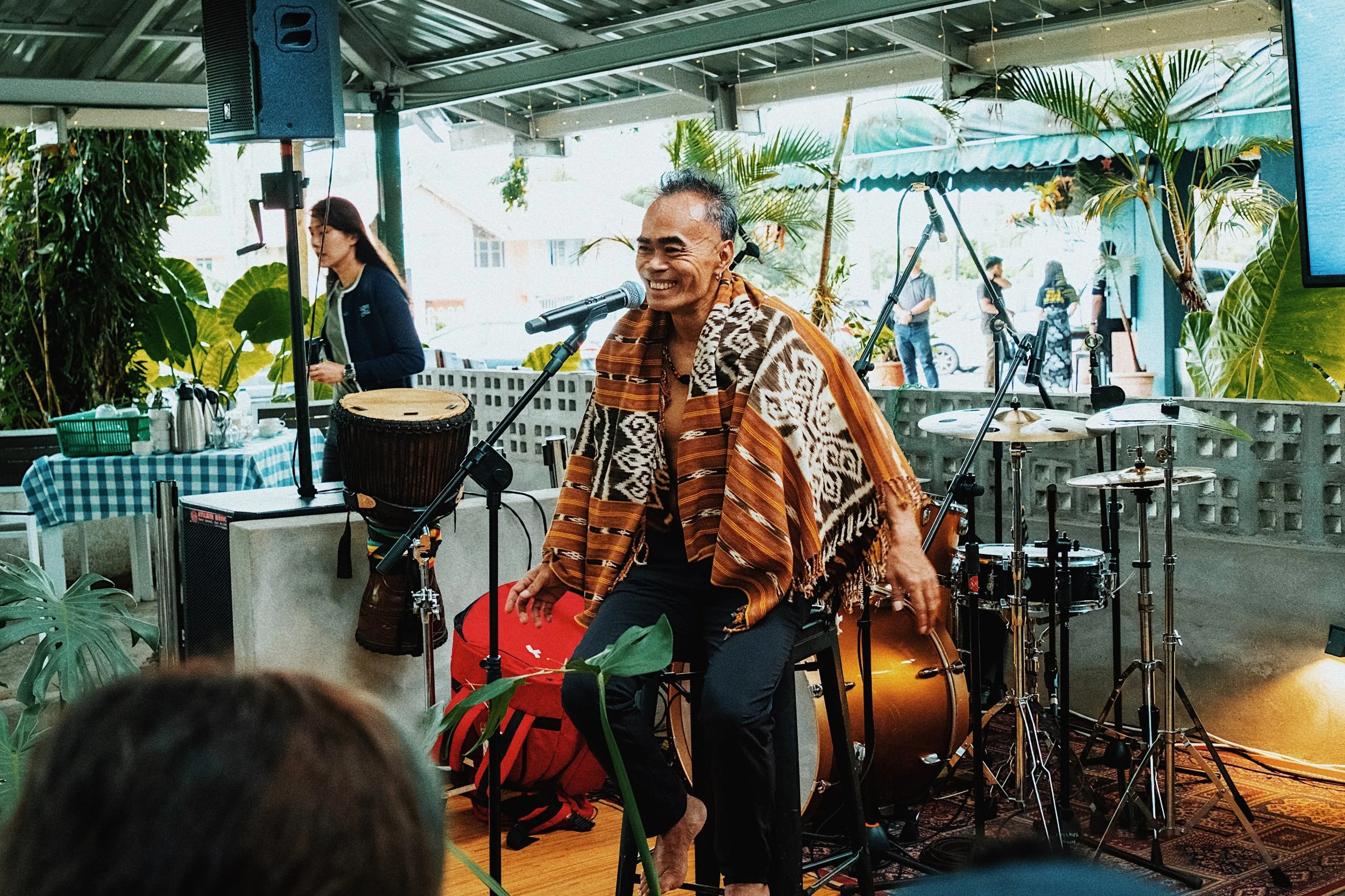A man sitting on a chair performing with a microphone, wearing a patterned shawl, in a lively indoor setting with musical instruments, plants, and people in the background.