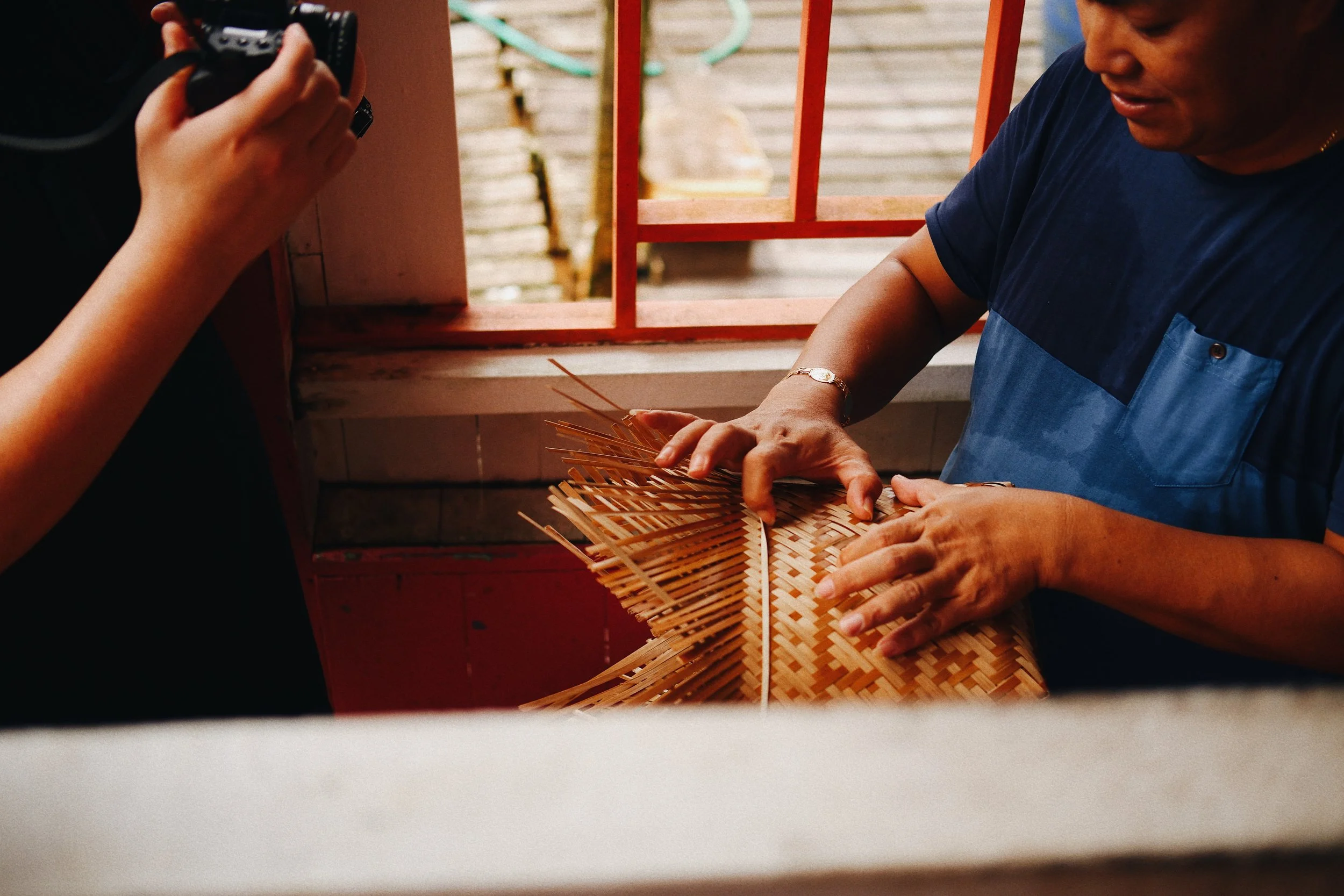 A woman demonstrates traditional weaving with bamboo strips while another person takes a photo of her.