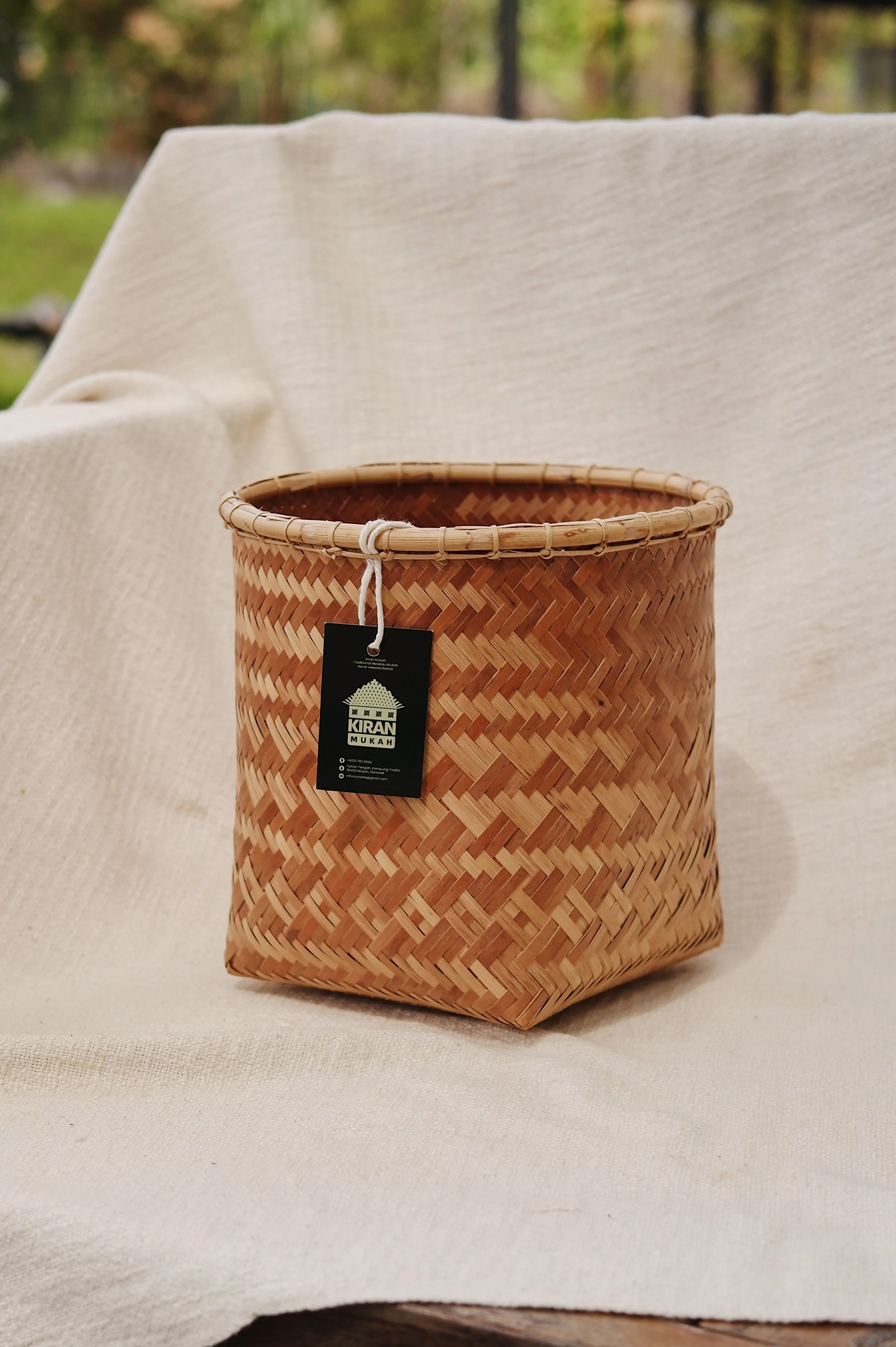 A woven wicker basket with a black tag attached, placed on a beige fabric surface with a blurred outdoor background of trees.