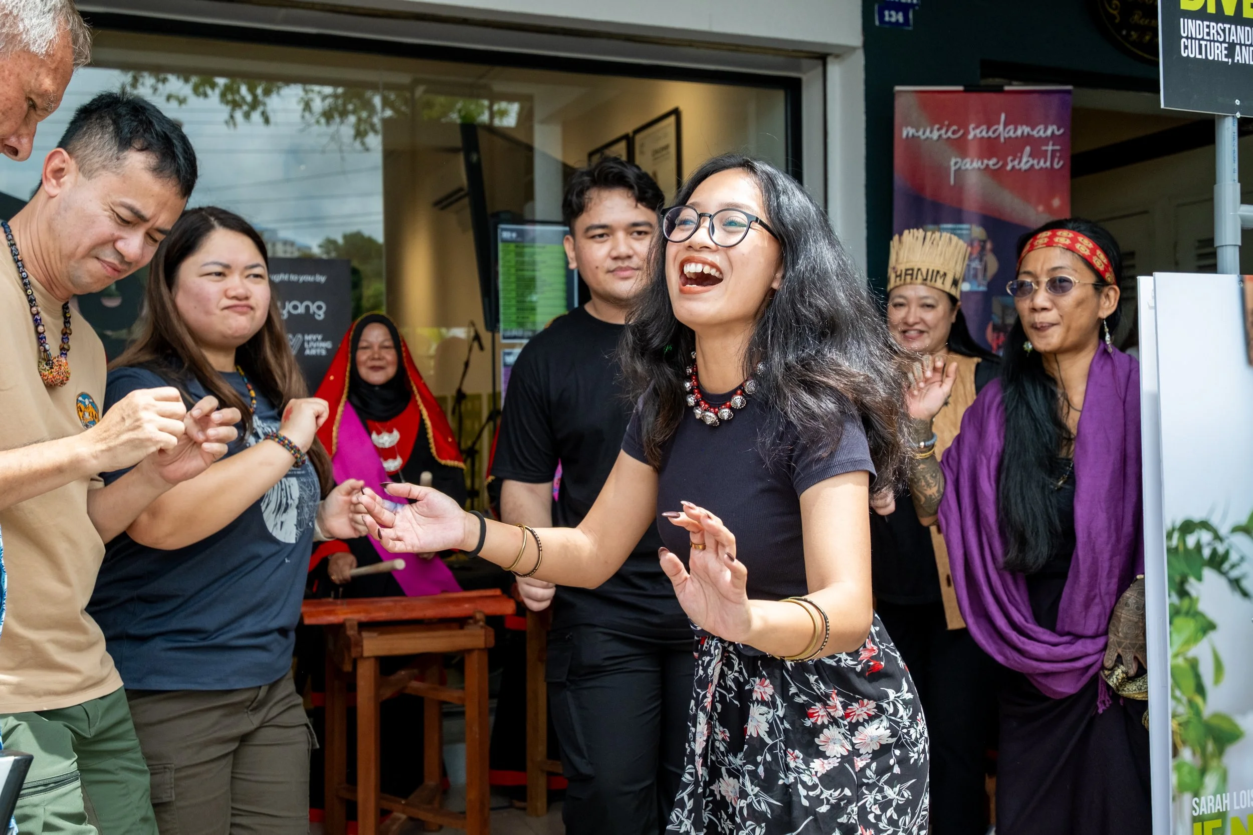 A group of people dancing and celebrating at an outdoor event, with an Asian woman smiling and holding hands with a man, surrounded by others who are dressed in colorful traditional and casual attire.