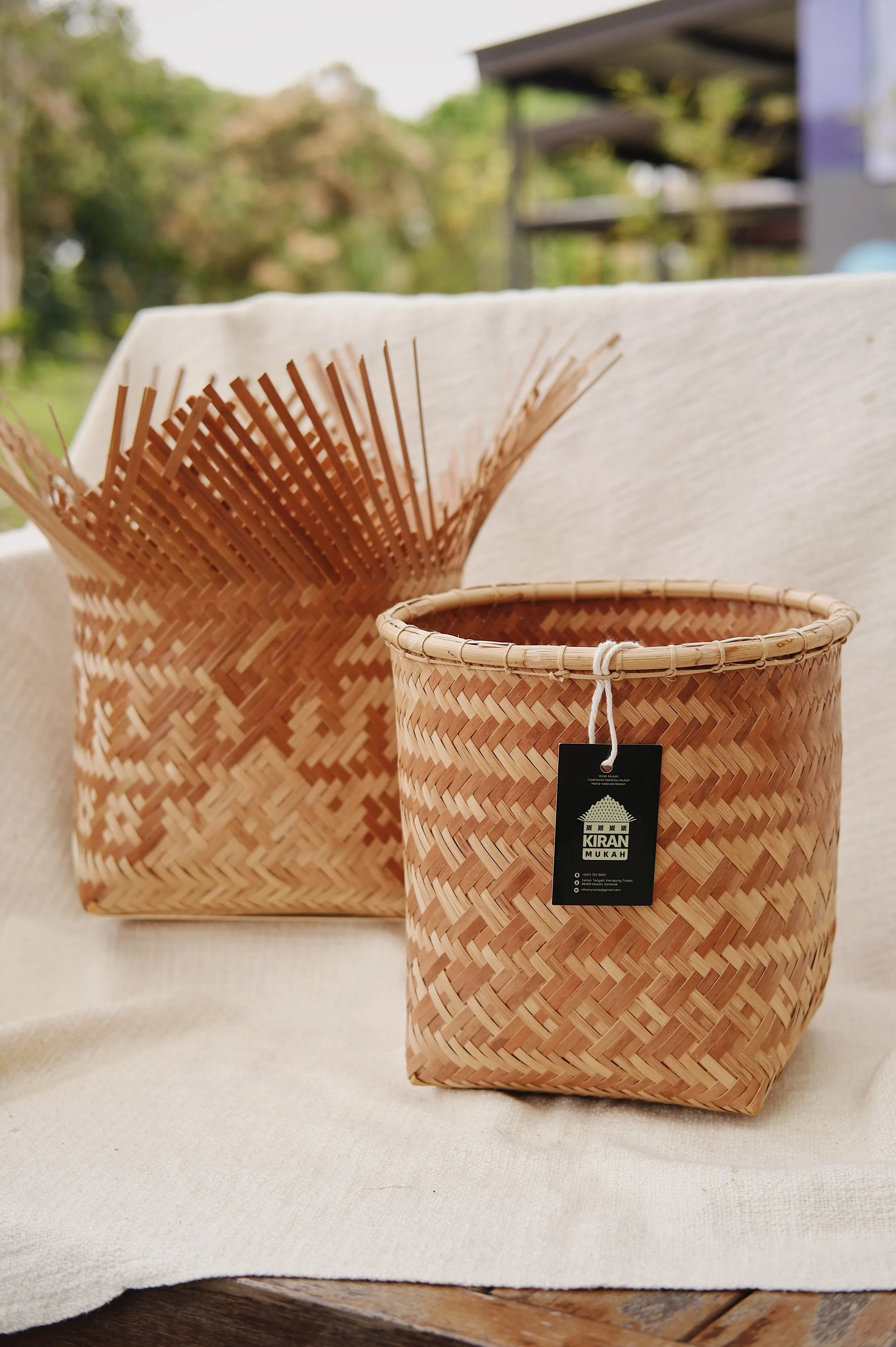 Two woven baskets, one larger and one smaller, displayed on a light-colored cloth with a blurred outdoor background.