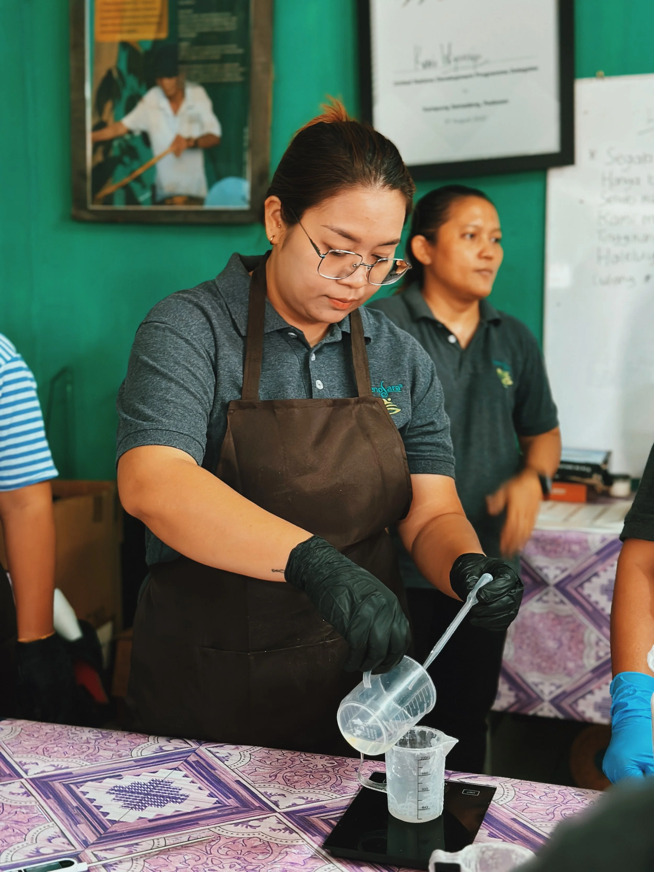 A woman wearing glasses, a gray polo shirt, a brown apron, and black gloves is pouring liquid into a measuring cup on a digital scale during a workshop or class.