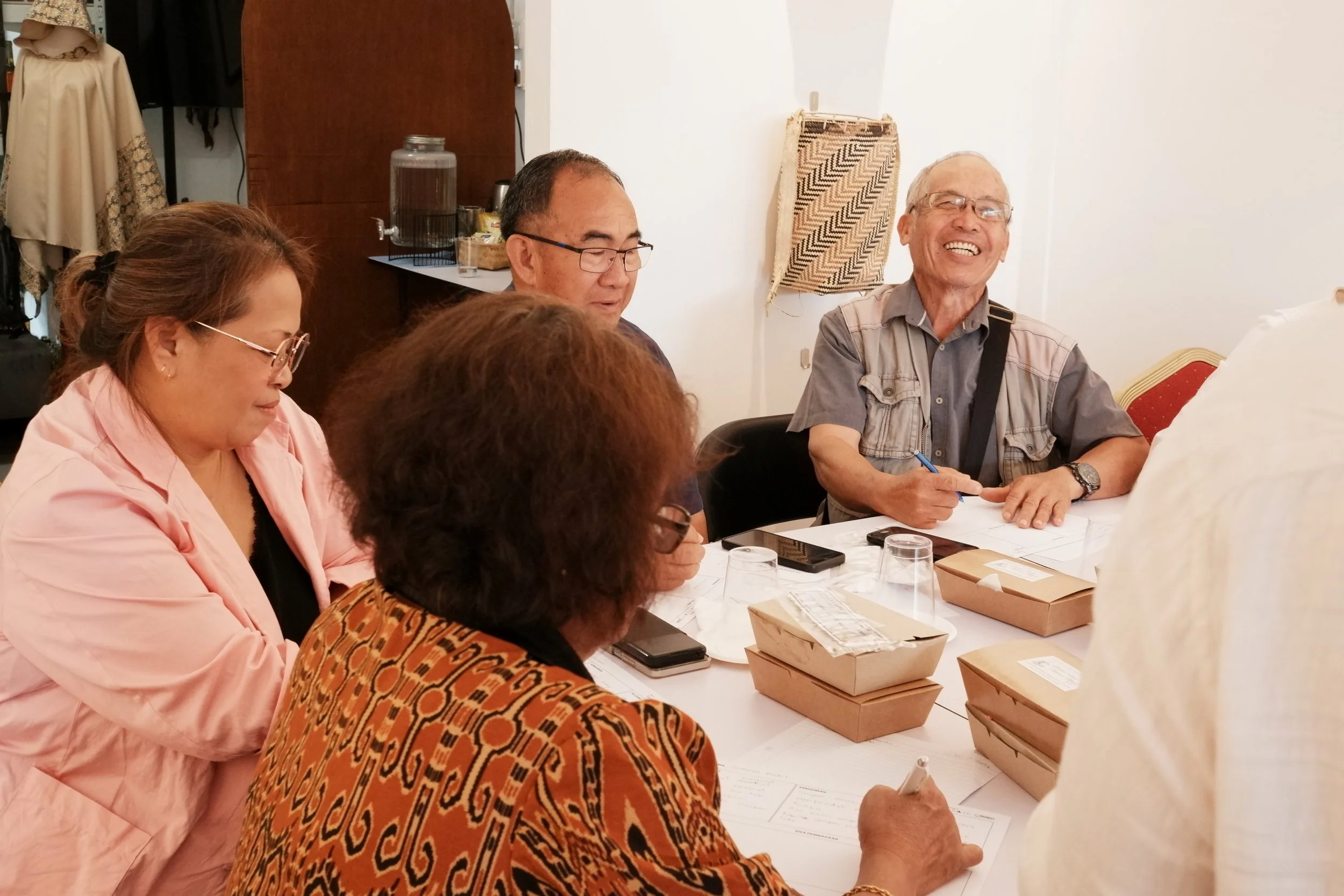 Group of five people sitting at a table, smiling and engaged in conversation, with papers, food containers, and drinks on the table.