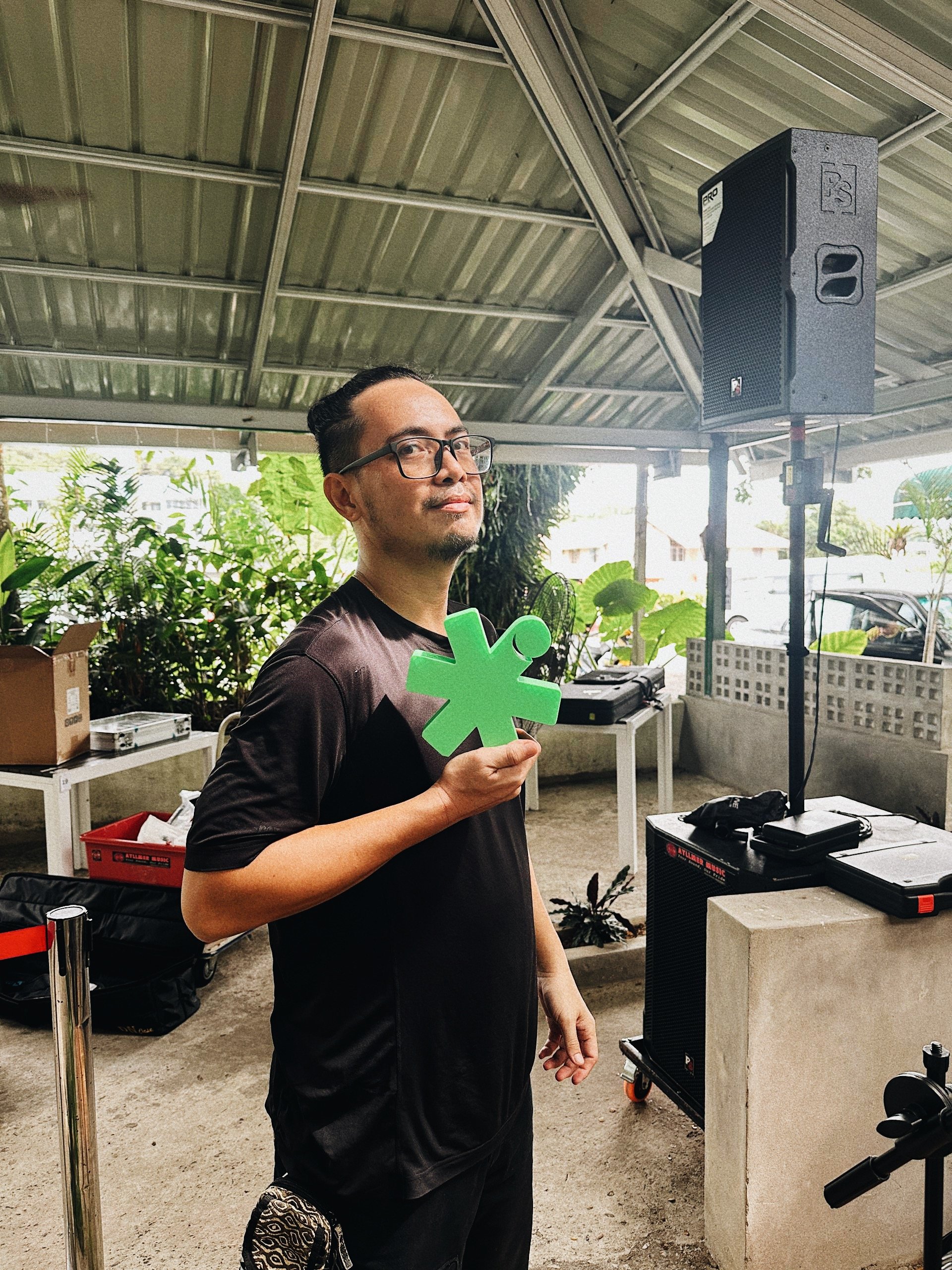 A man with glasses holding a green asterisk symbols in an outdoor covered area with plants and audio equipment.