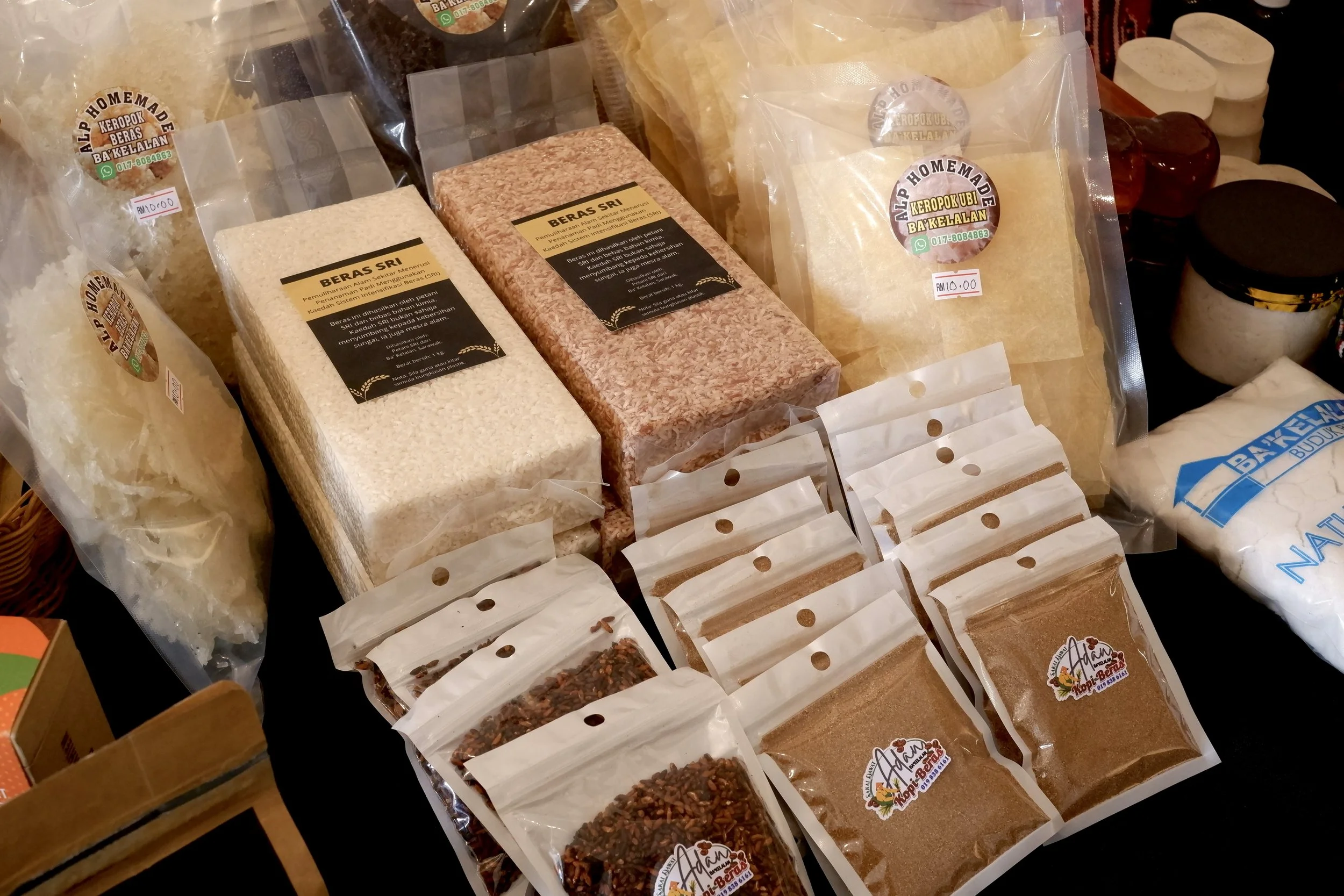 Various packaged rice products, including white rice, red rice, and rice flour, on a market table with labels and prices, along with other food items in the background.