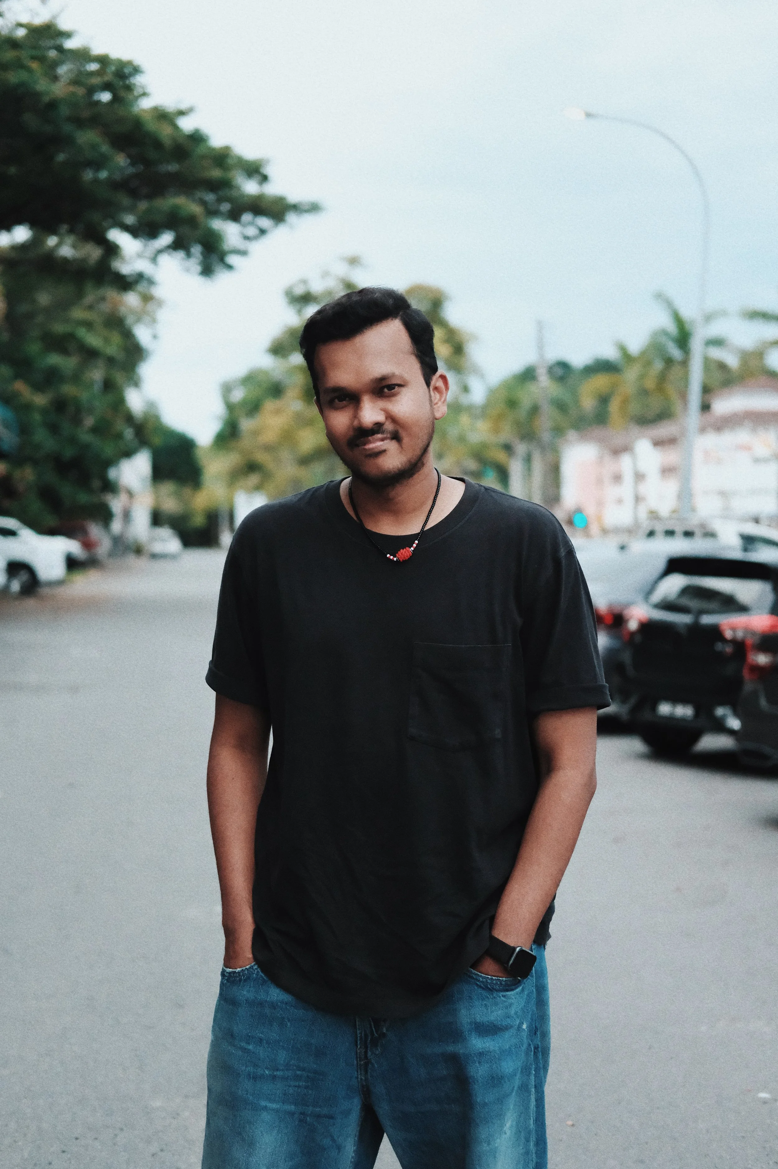 A young man in a black t-shirt and jeans standing on a street with parked cars and trees in the background.