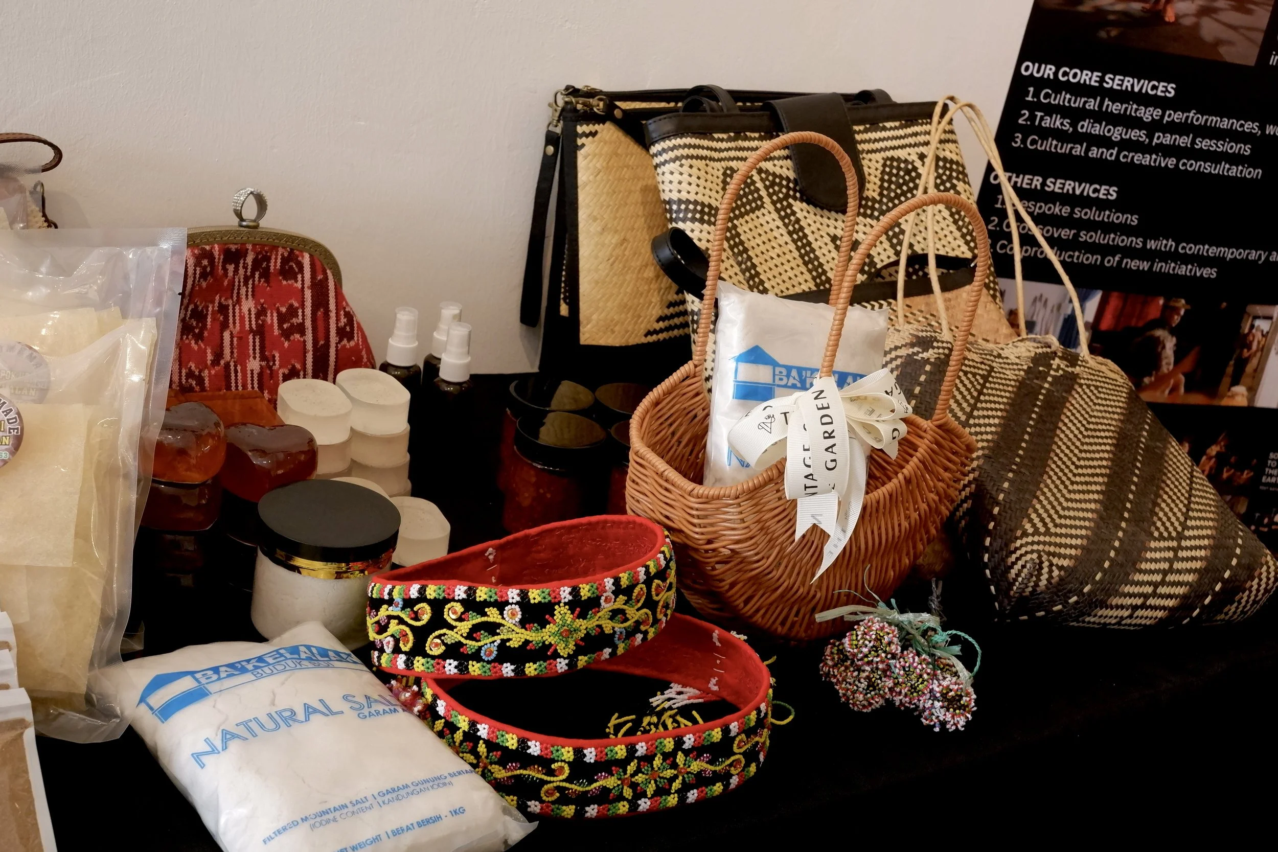 A table displays various handmade crafts and natural products, including beaded headbands, woven bags, jars of salt, soap, and skincare items, along with a poster detailing core and other services.