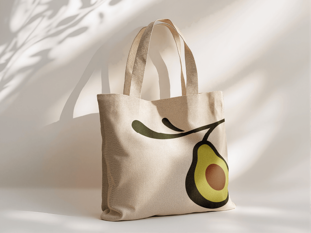 Beige tote bag with an illustration of an avocado on it, set against a white background with shadows.