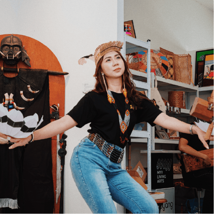 Young woman wearing a Native American-inspired headdress and jewelry, dancing or performing in a room decorated with Native American artifacts and artwork.