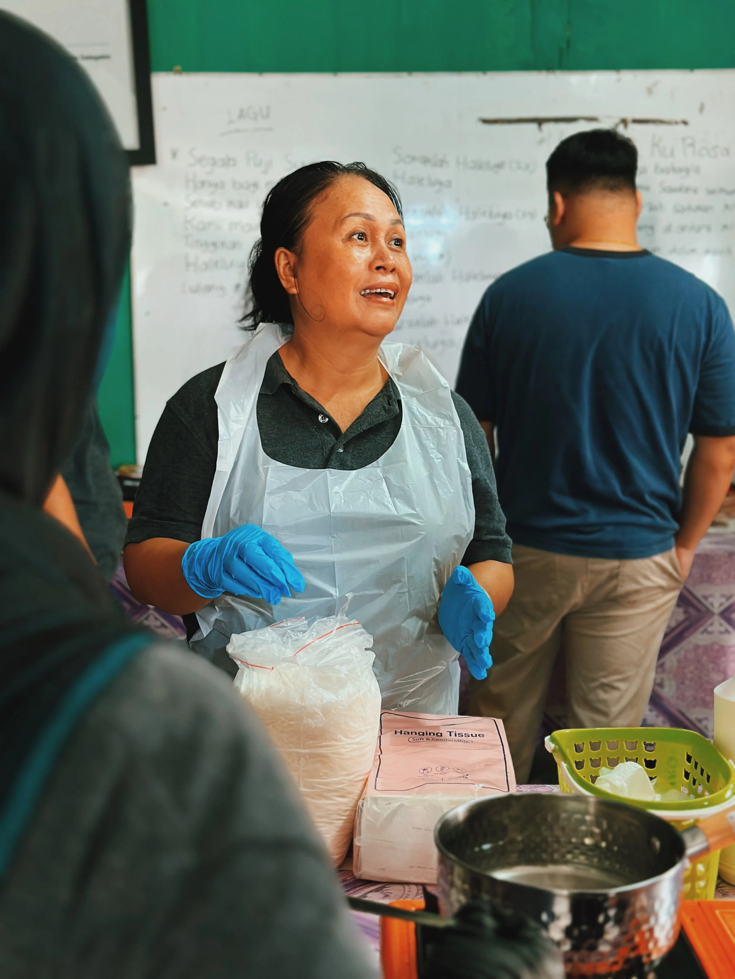 A woman with black hair wearing a gray shirt, a plastic apron, and blue gloves is talking to a group of people in a classroom setting. There are cooking supplies and a whiteboard in the background.