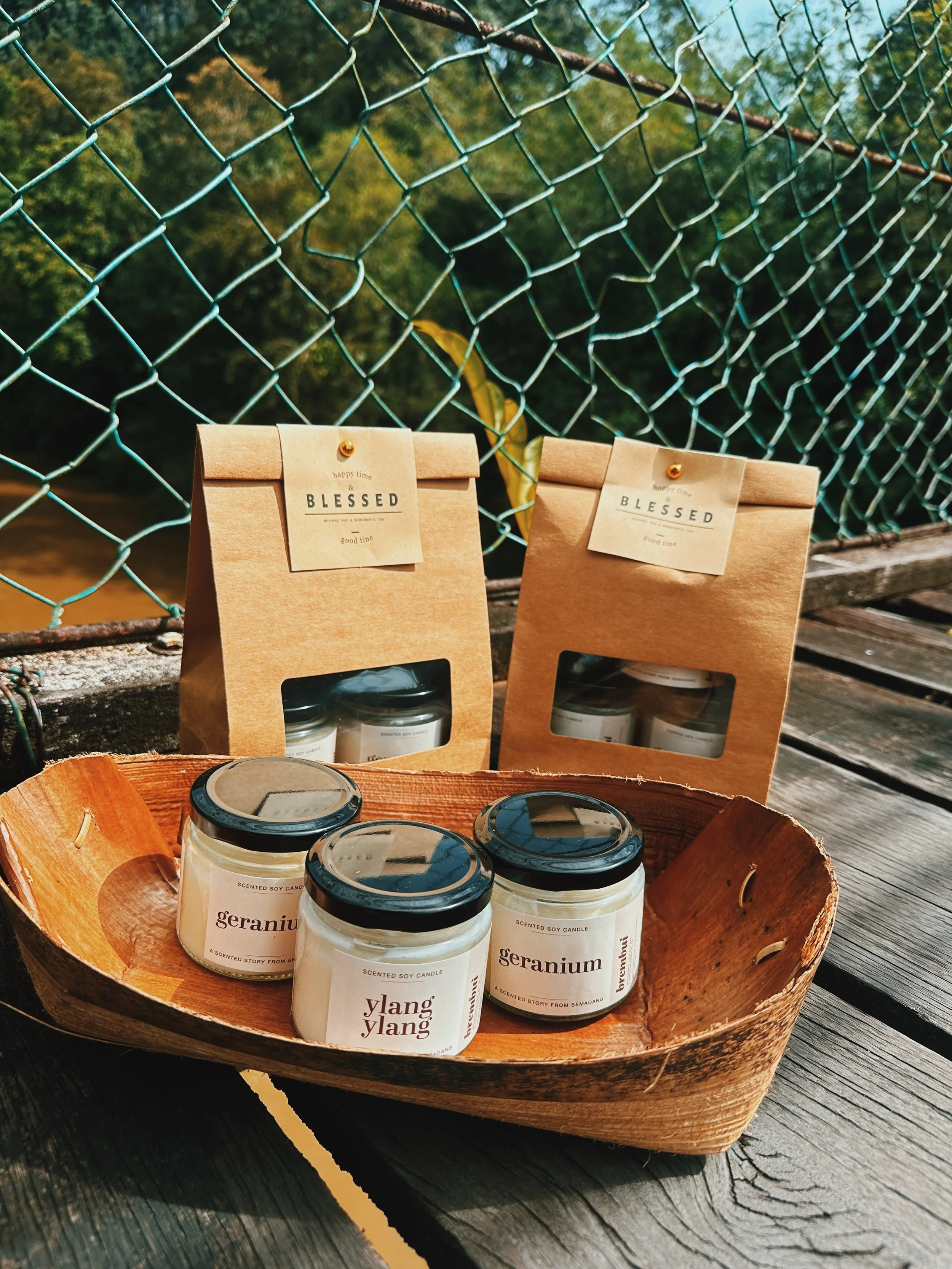 Wooden tray holding three scented candles with labels: geranium and ylang ylang, placed outdoors on a weathered wooden surface, with two paper bags labeled 'Blessed' in the background, against a chain-link fence and green foliage.