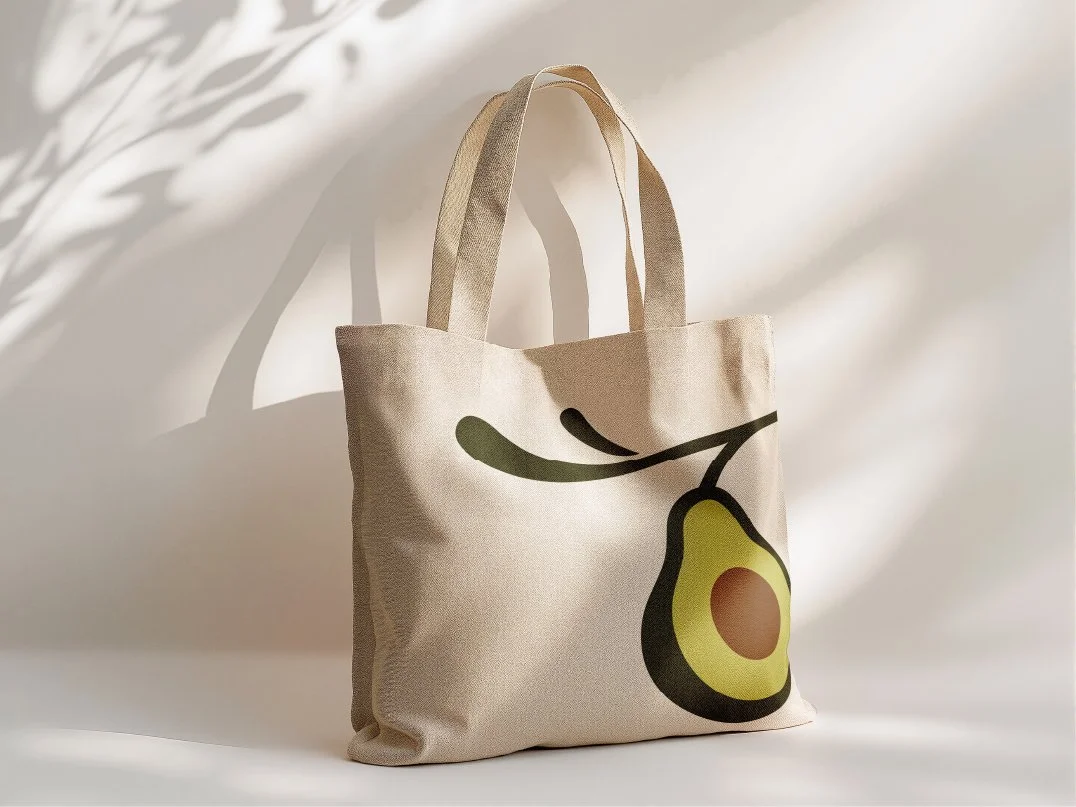 A beige tote bag with an avocado graphic design, placed against a light background with shadows of leaves.